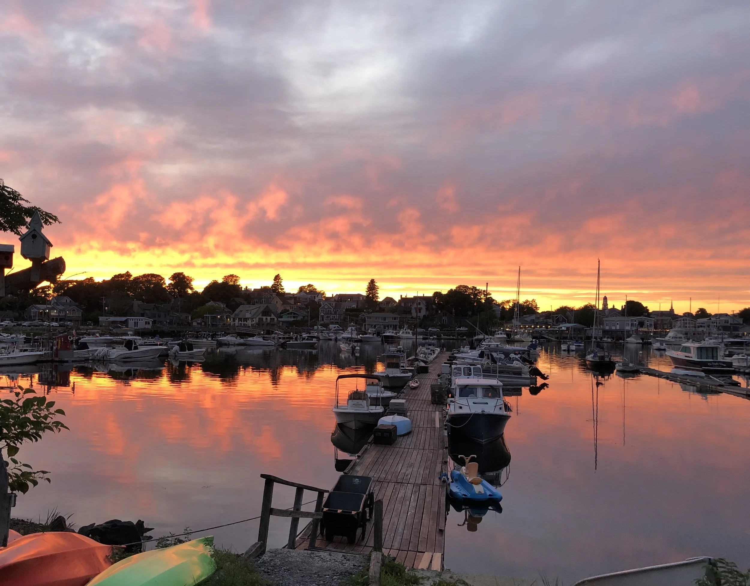 A marina at sunset with orange and pink clouds reflected in the water, boats docked along a wooden pier, and houses in the background.