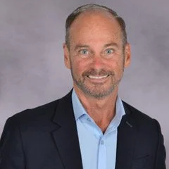 Professional headshot of a middle-aged man with short, light brown hair and a beard, wearing a dark blazer and a light blue shirt, smiling against a gray background.
