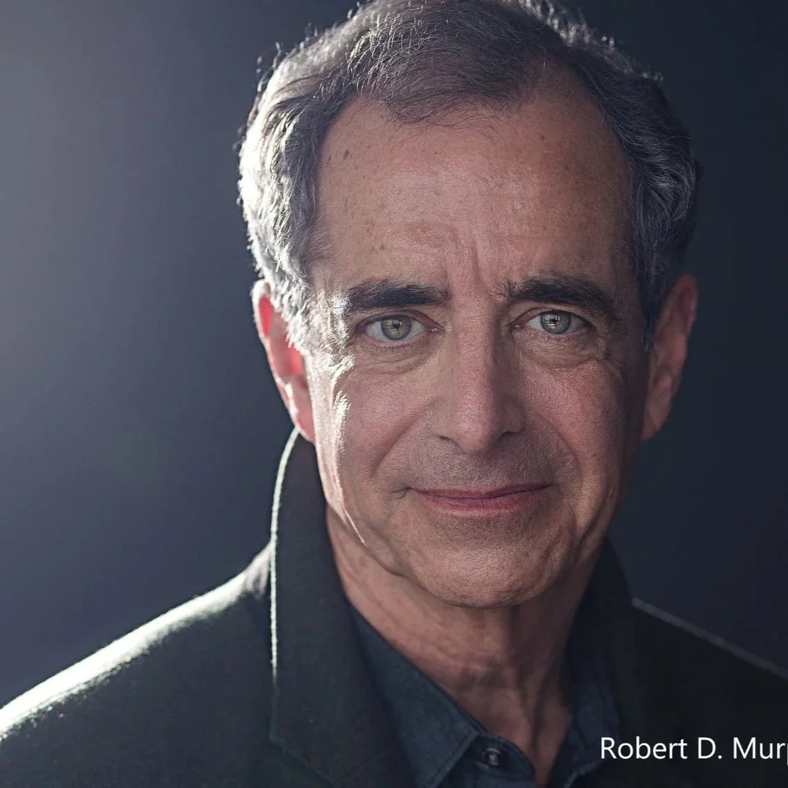 Close-up portrait of a middle-aged man with gray hair and blue eyes, wearing a dark shirt and jacket, against a plain background.