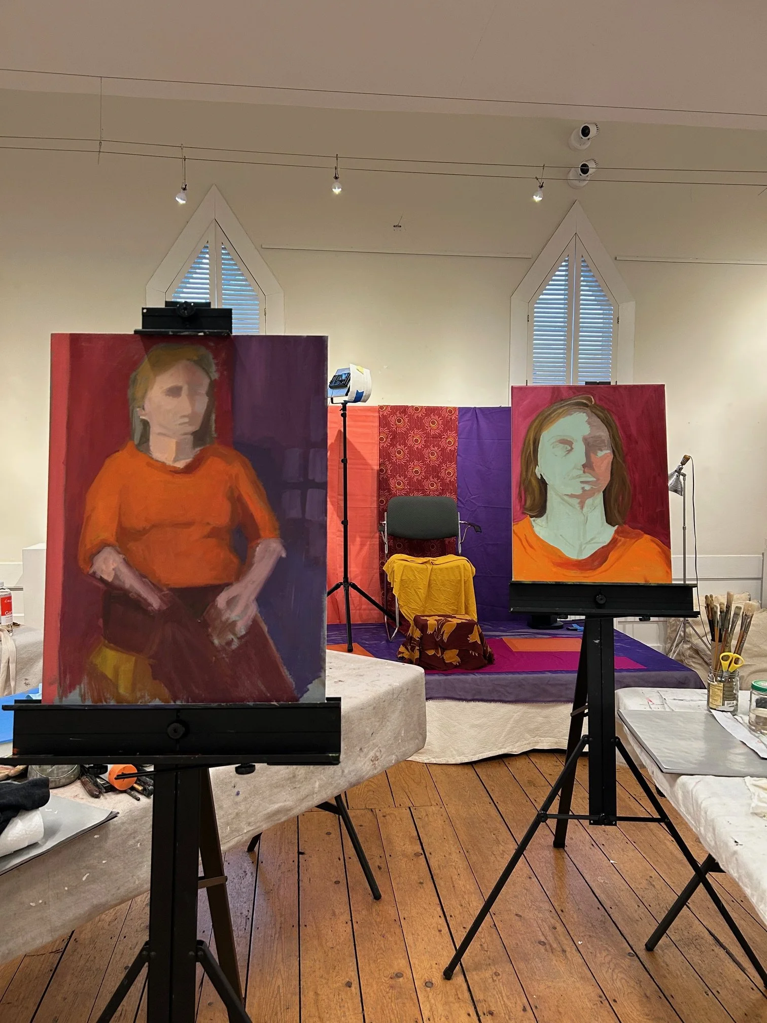 An art studio with two portrait paintings on easels, a stage with a yellow cloth-covered table and microphone, and a backdrop of colorful fabric.