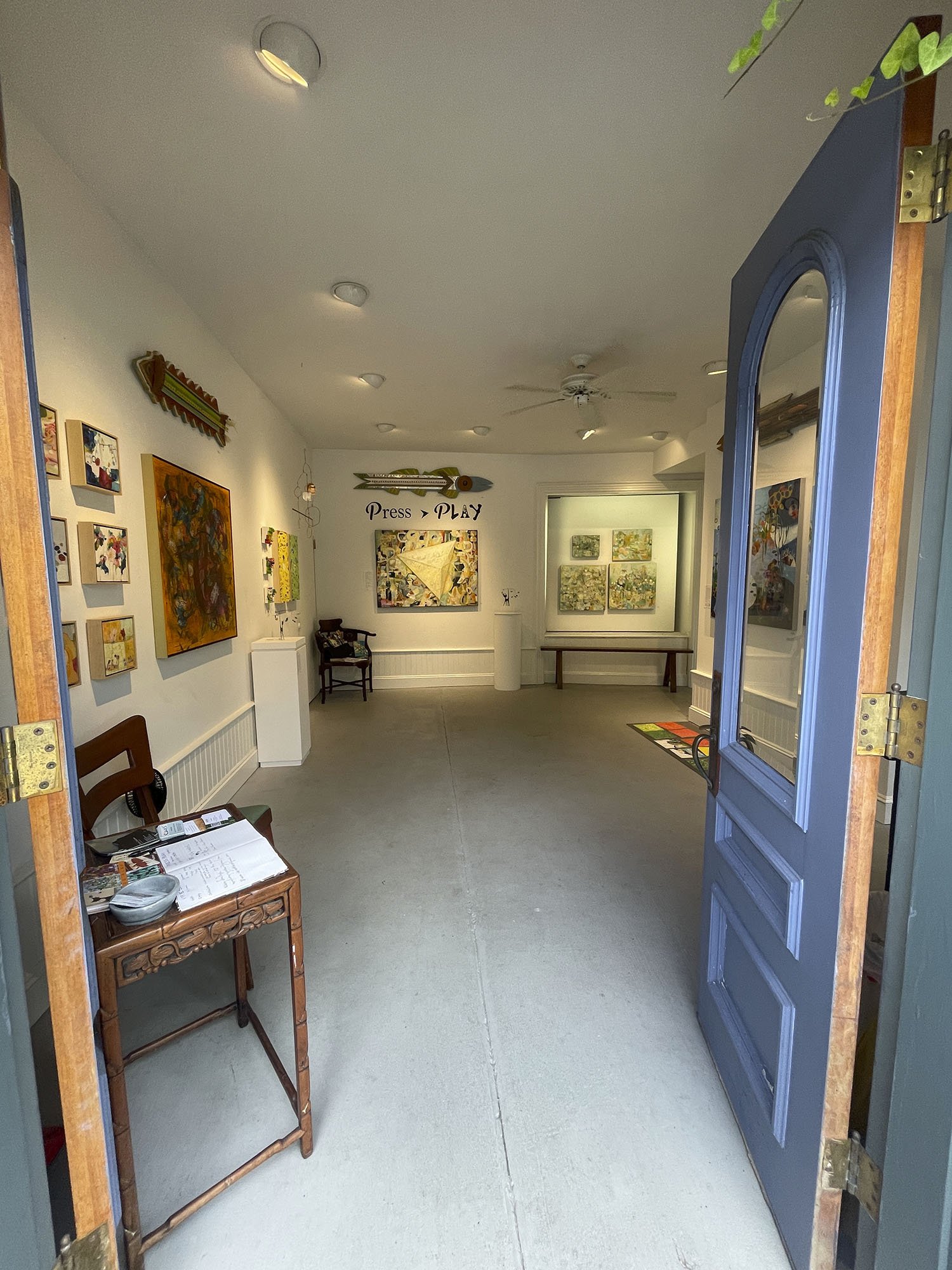 Art gallery interior with various paintings, a blue door to the right, a small table with papers, and a 'Press Play' sign on the wall.