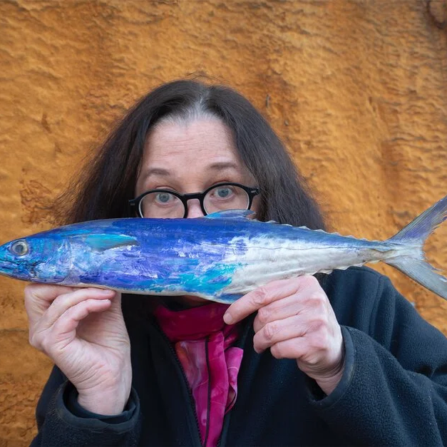 Woman with glasses holding a painted fish model in front of her face, standing against an orange wall.