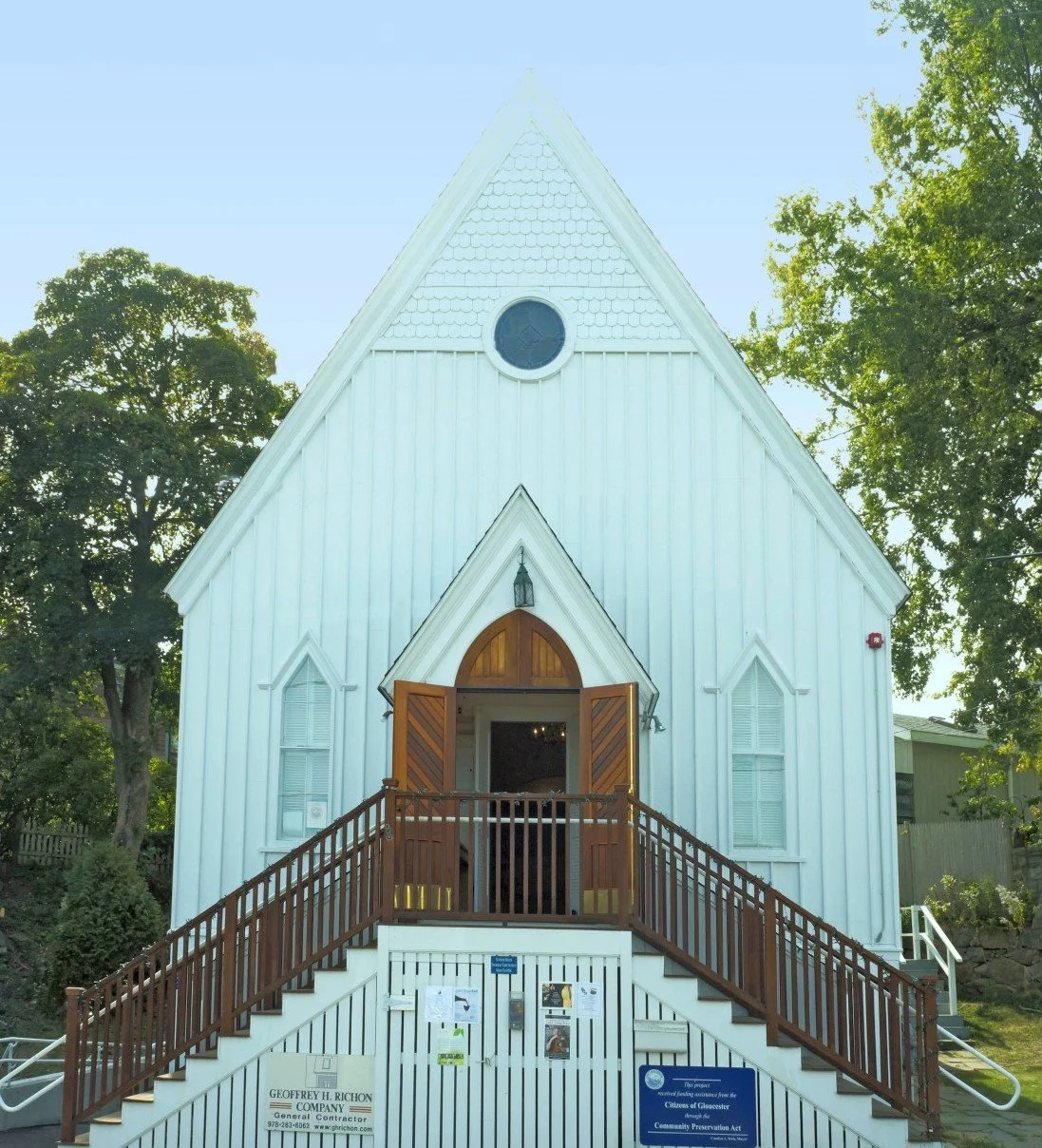 Small white church with a wooden front door, front staircase, and steep roof, surrounded by trees.