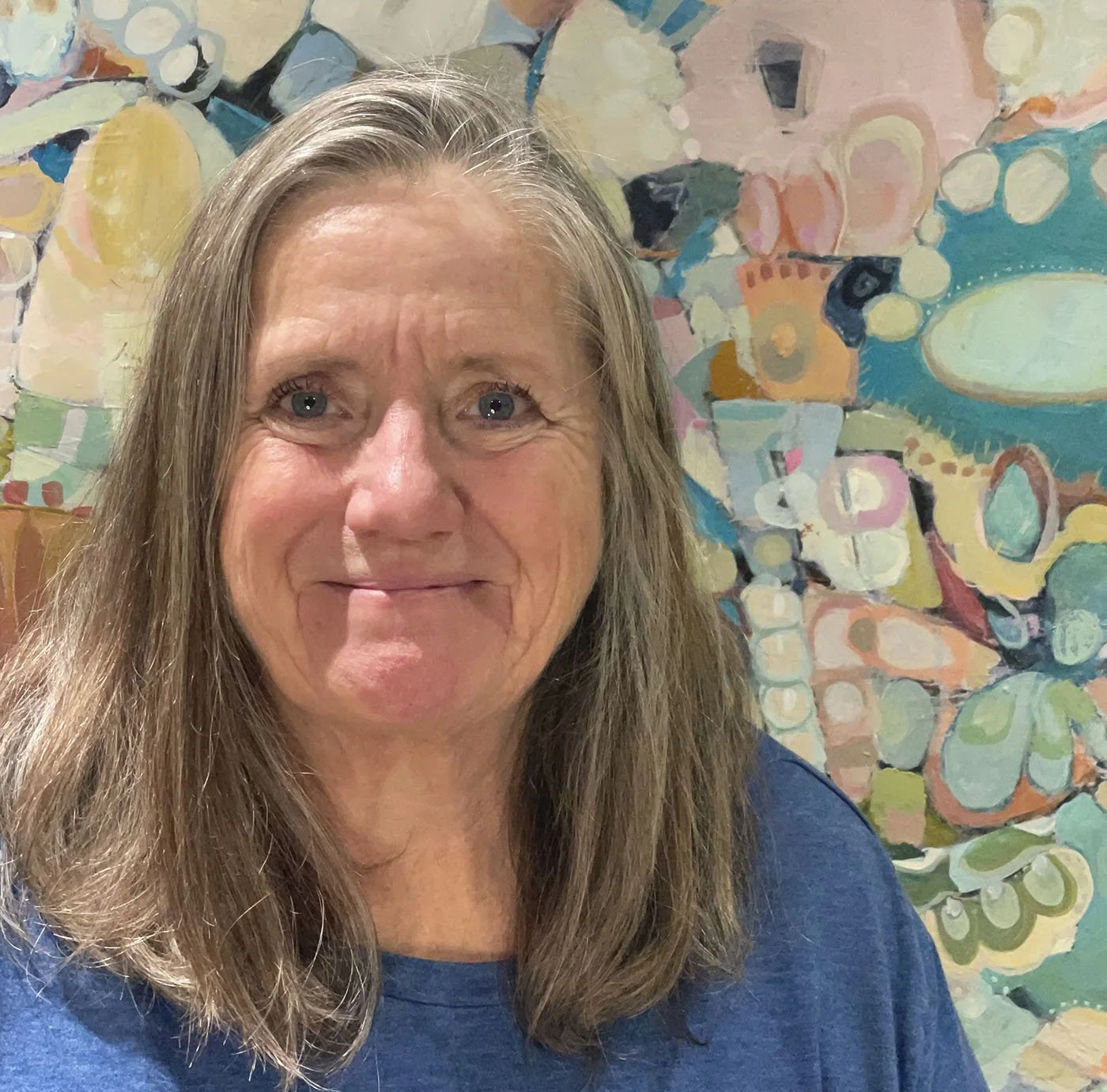 A smiling woman with long, light brown hair standing in front of an abstract colorful painting.