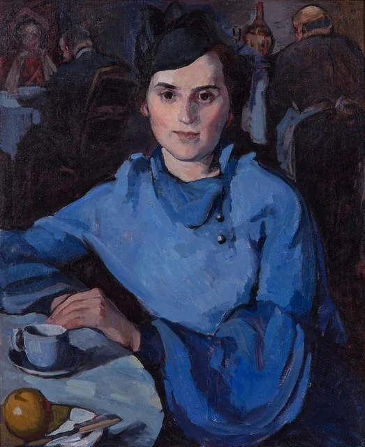 A woman with dark hair wearing a blue dress sits at a table with a cup and napkin, in a restaurant scene with other diners in the background.
