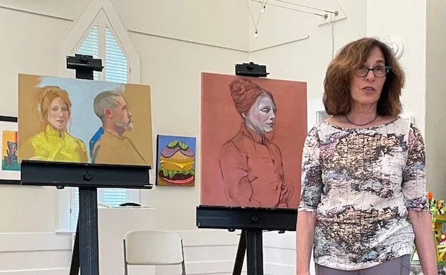 A woman standing inside an art gallery with her artwork on display behind her, including a colorful portrait of a woman with red hair, a painting of a landscape with a rainbow and mountains, and a side-profile of a woman and man in a classic portrait style.