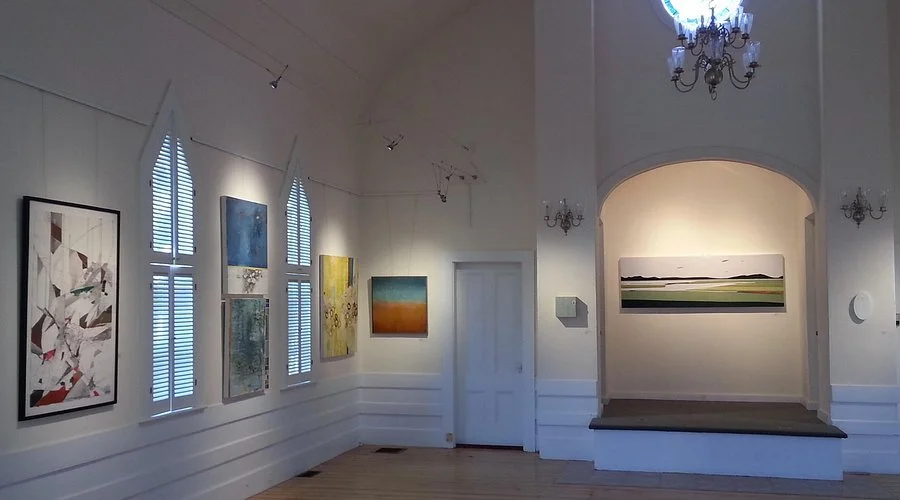 An art gallery with paintings on the walls, arched windows with shutters, chandeliers, and minimalist interior design.