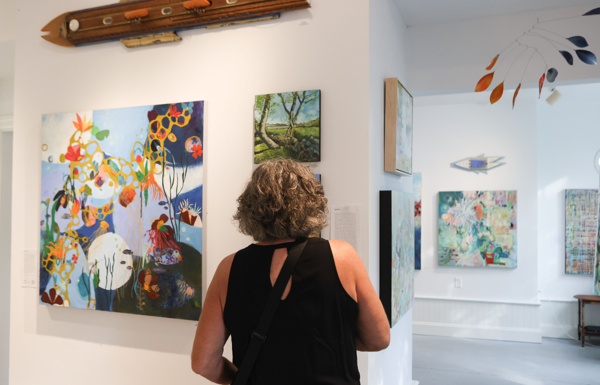 A woman with curly gray hair wearing a black sleeveless top is viewing abstract and landscape paintings in an art gallery with white walls.