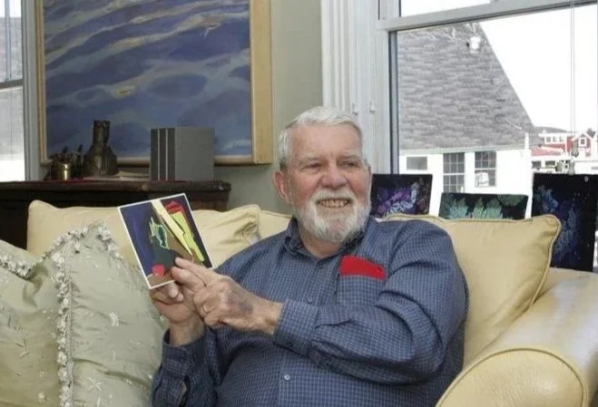 An elderly man with a beard and white hair sitting on a beige couch, holding a colorful greeting card and smiling. The room has a window with a view of houses outside, a painting of a seascape on the wall, and multiple computer monitors in the background.