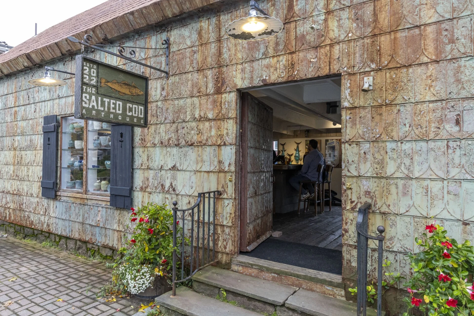 Exterior view of The Salted Cod Art House restaurant with rustic brick walls, a hanging sign, and window shutters. The entrance has a small step with black metal railings, and a man is sitting at the bar inside visible through the open doorway, with sea-themed decor on the walls.