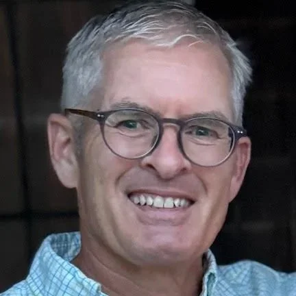 Close-up of a smiling middle-aged man with gray hair, wearing glasses and a blue checkered shirt.