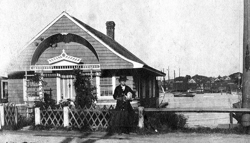 A woman standing in front of a small house, holding an object, in a black and white photo from a past era.