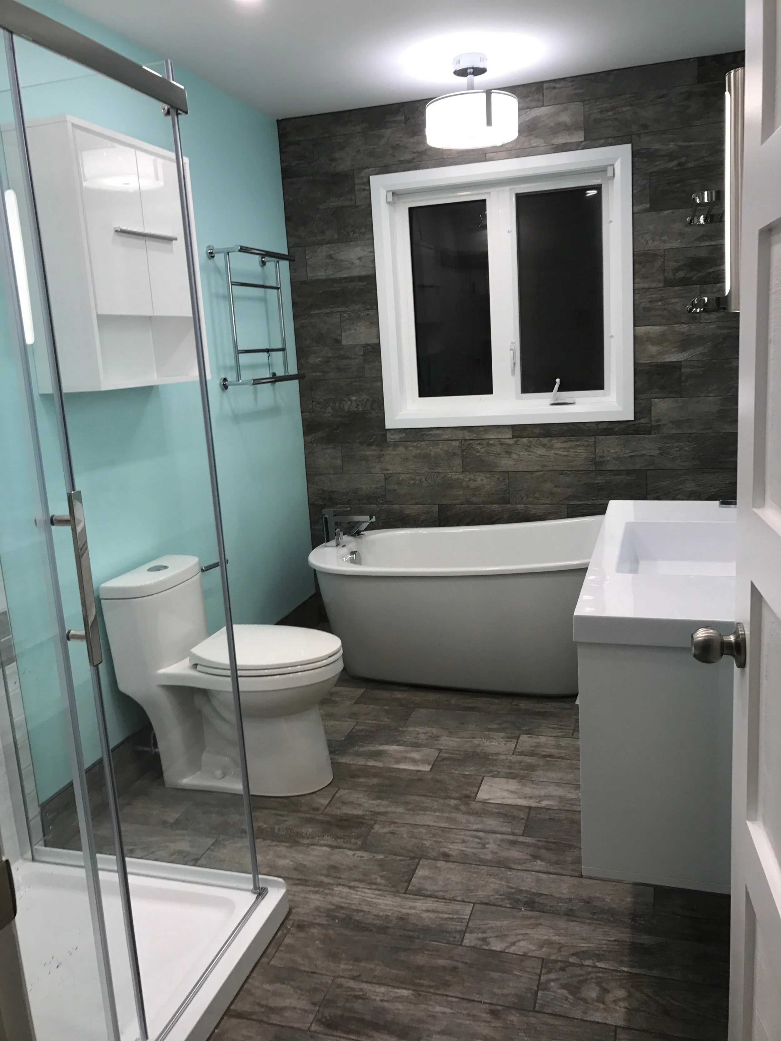 A modern bathroom with a shower, toilet, bathtub, and sink. Blue and wood-textured wall tiles, a window, and contemporary lighting fixtures.