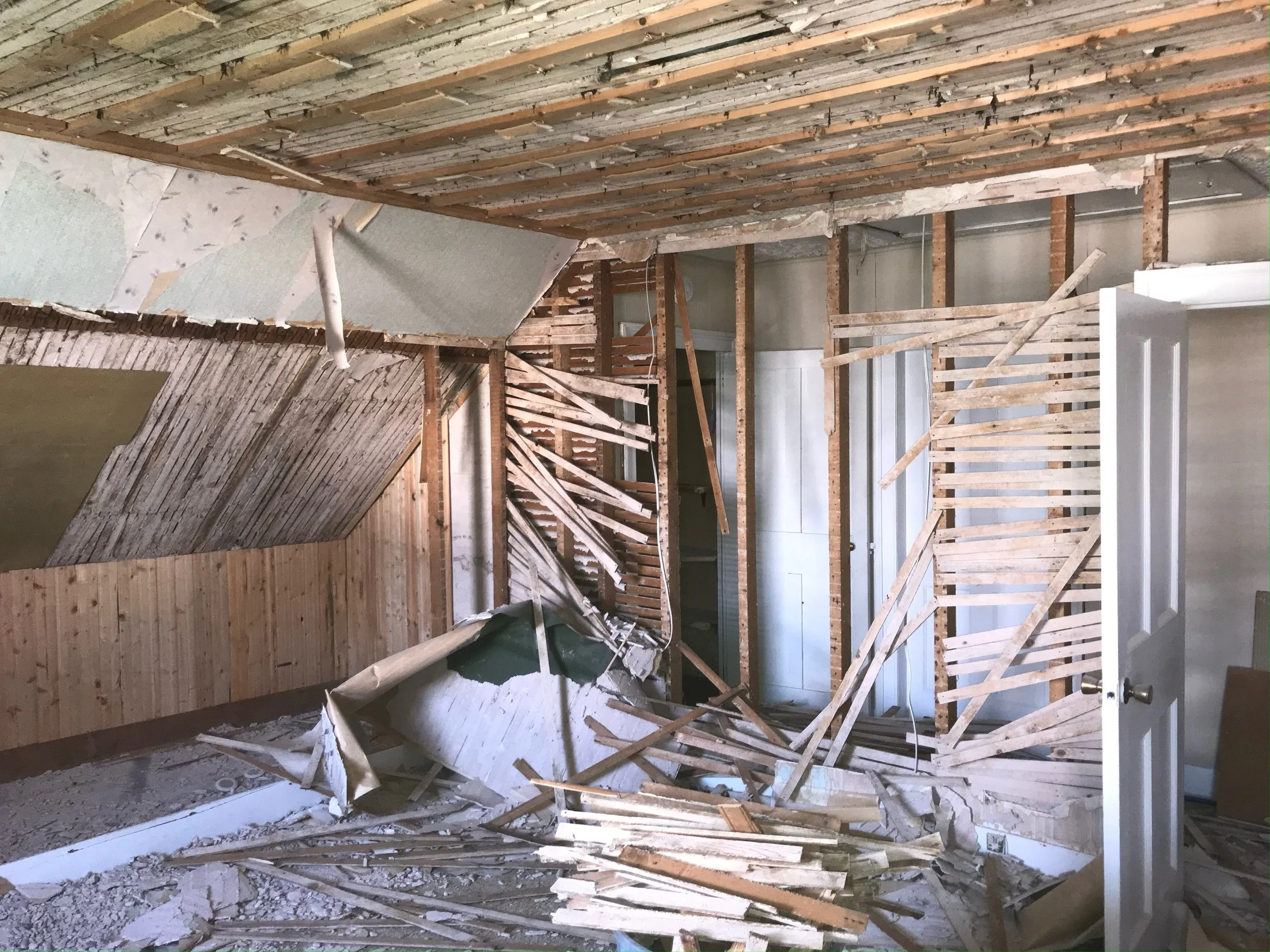 Interior of a room under renovation with damaged walls and ceiling, exposed wooden studs, and debris on the floor.