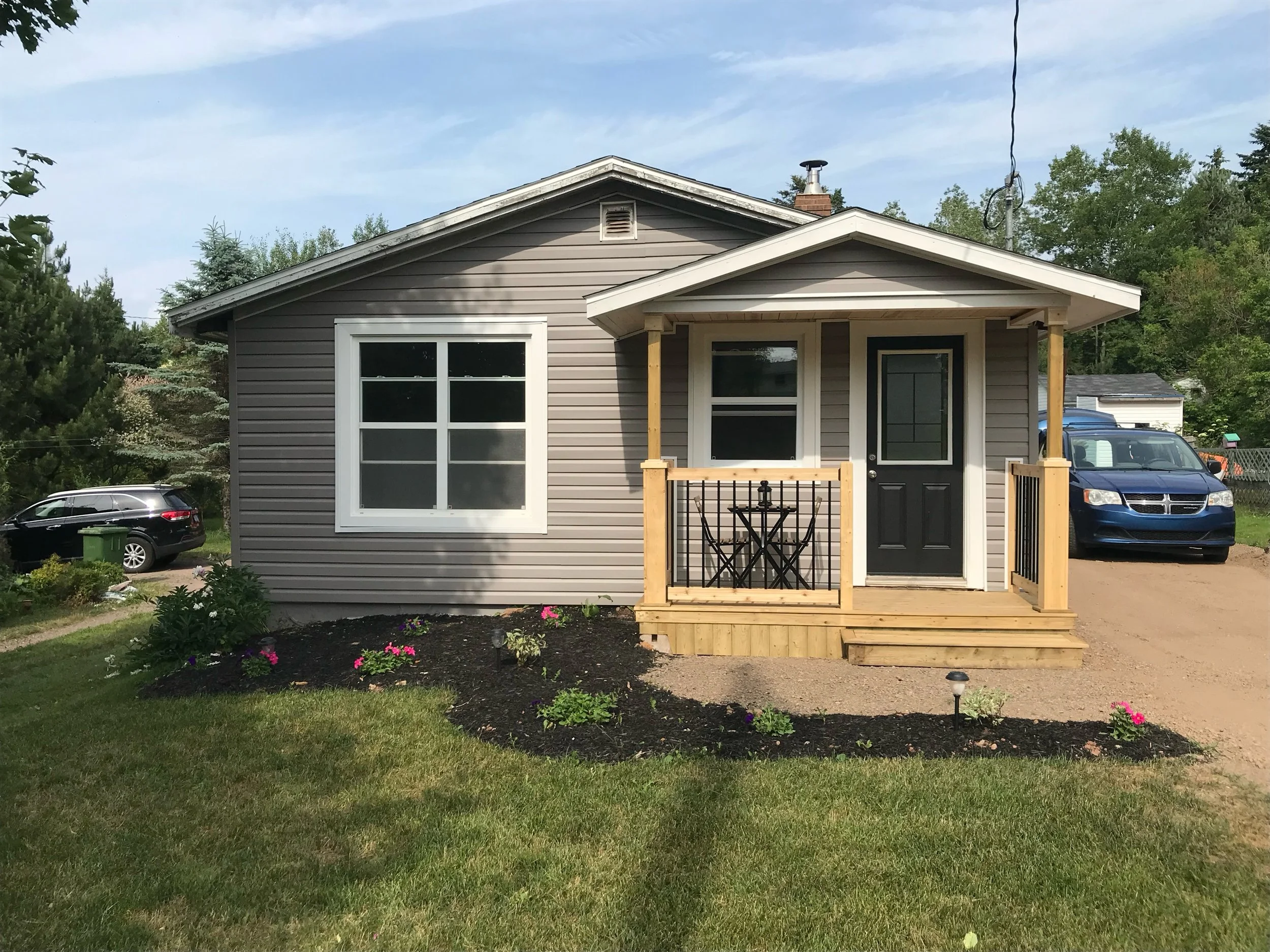 A house with gray siding, a small front porch with wooden steps, and a black door. Two windows on the front, a landscaped yard with flowers, and cars parked in the driveway.