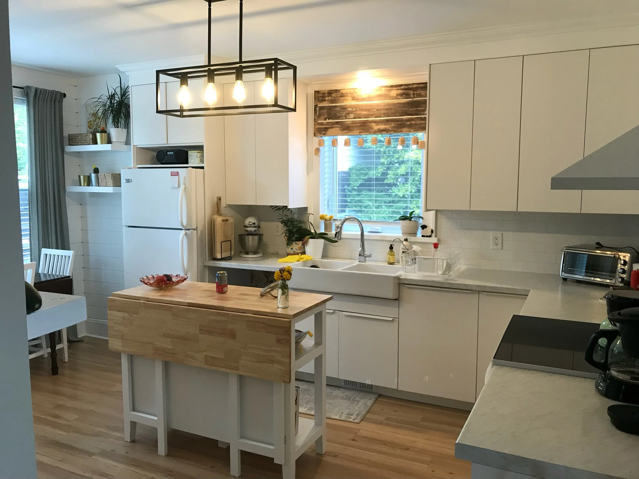 Bright kitchen with white cabinets, a wooden island, and a farmhouse sink under a window. Decor includes plants, a light fixture, and various kitchen appliances.