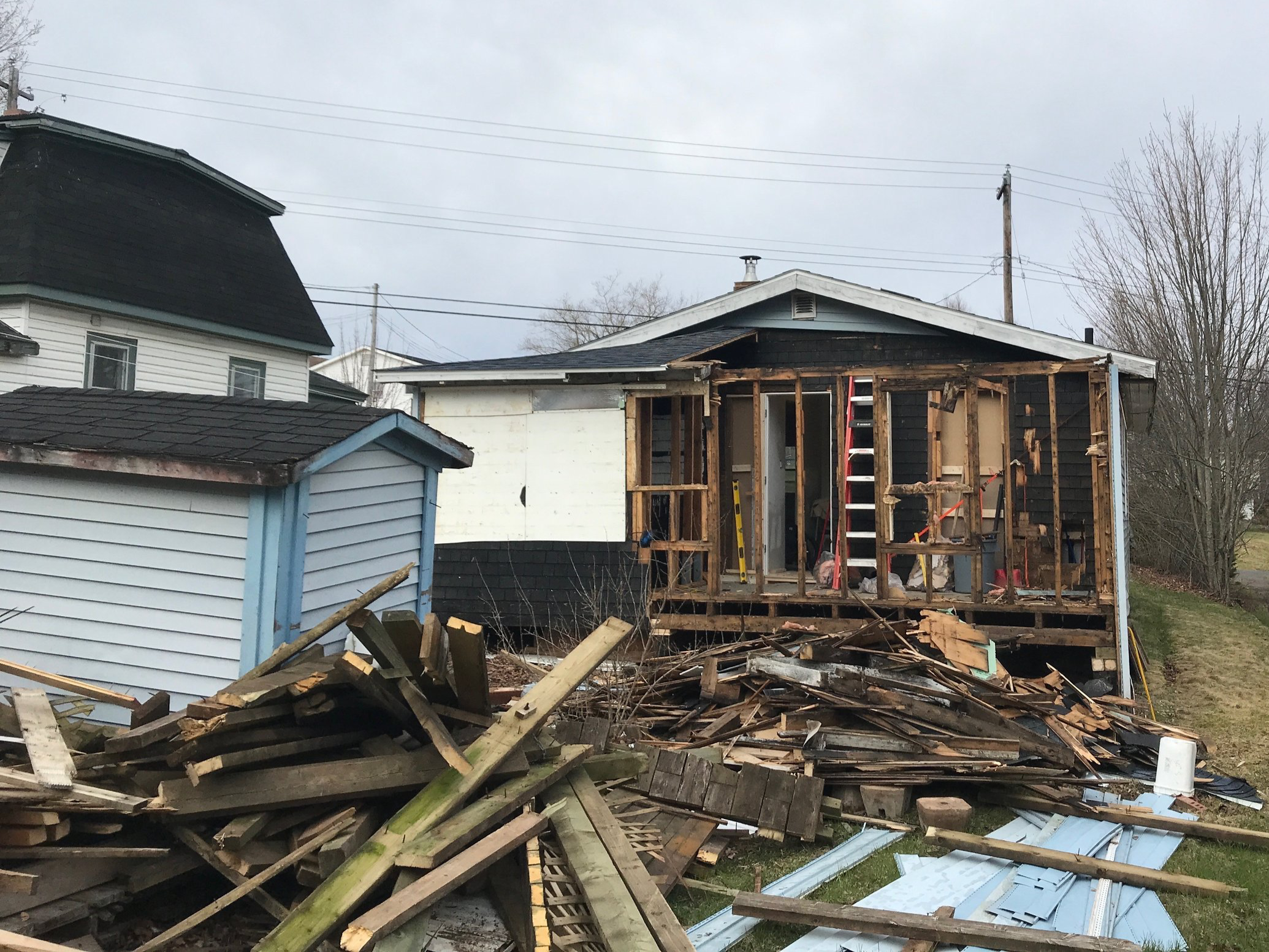 House under renovation with exposed wooden framing, scattered construction debris, and roof damage, neighboring houses visible in background.
