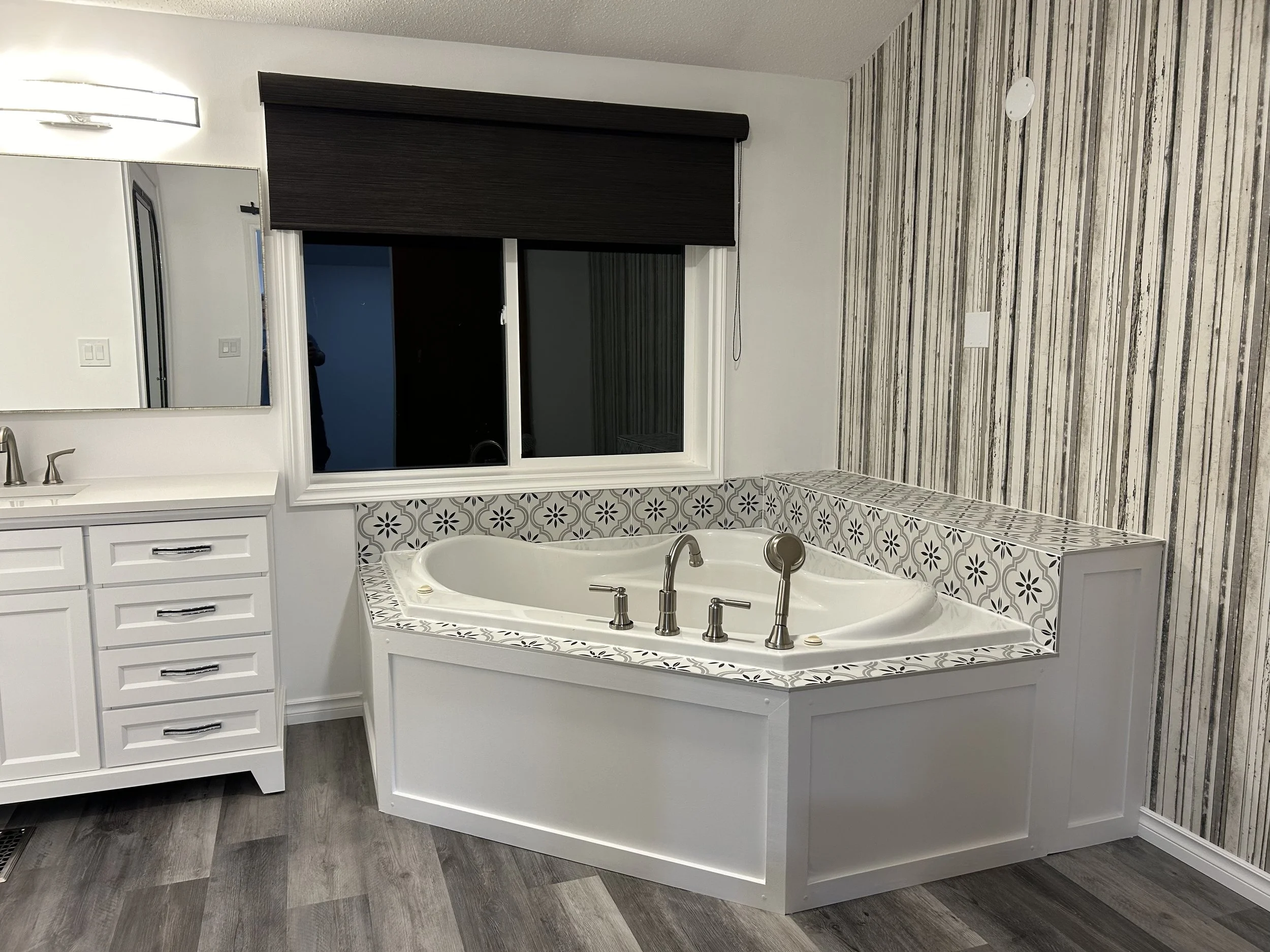 Bathroom with a large bathtub surrounded by decorative black and white tiles, white vanity with cabinet drawers, mirror, and faucet, window with a dark blind, and wood-patterned wall paneling.