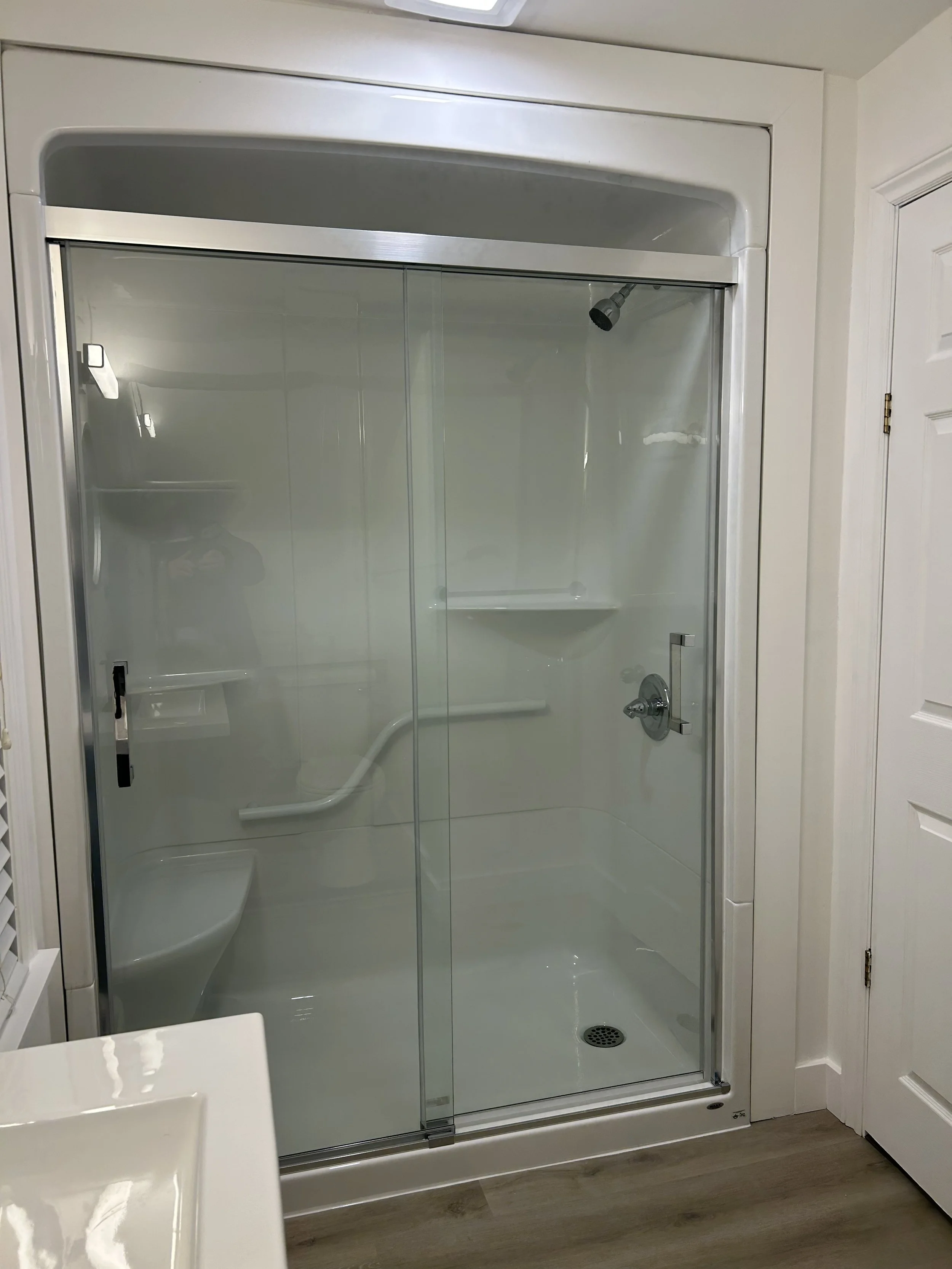 A walk-in shower with glass sliding doors, white interior walls, a showerhead, a control handle, built-in shelves, a safety grab bar, and a drain on the floor. Part of a white door and a bathroom wall are visible.