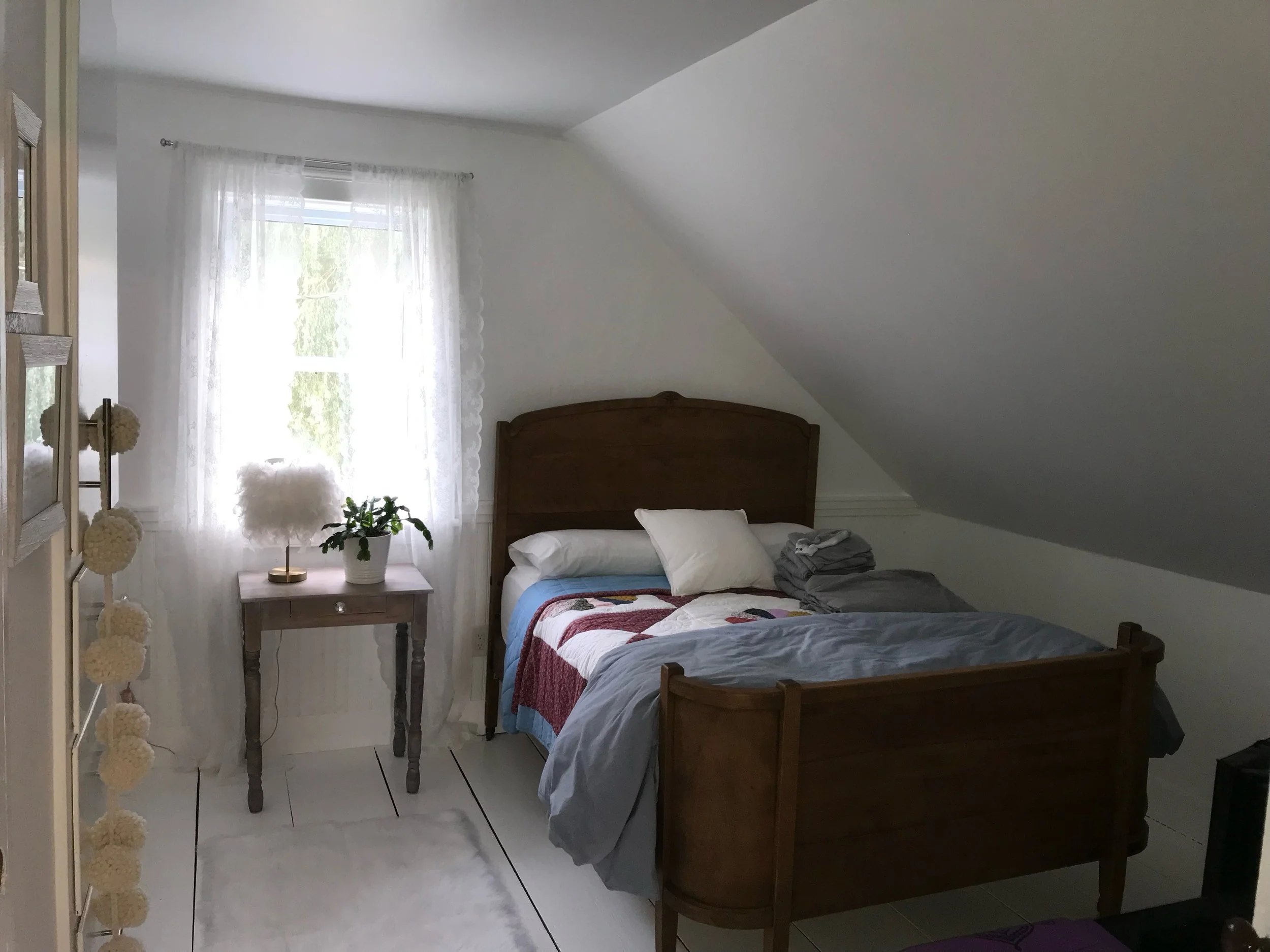 A cozy bedroom with a wooden bed, white pillows, and a comforter, next to a small nightstand with a plant and a lamp, illuminated by natural light from a window with white curtains.