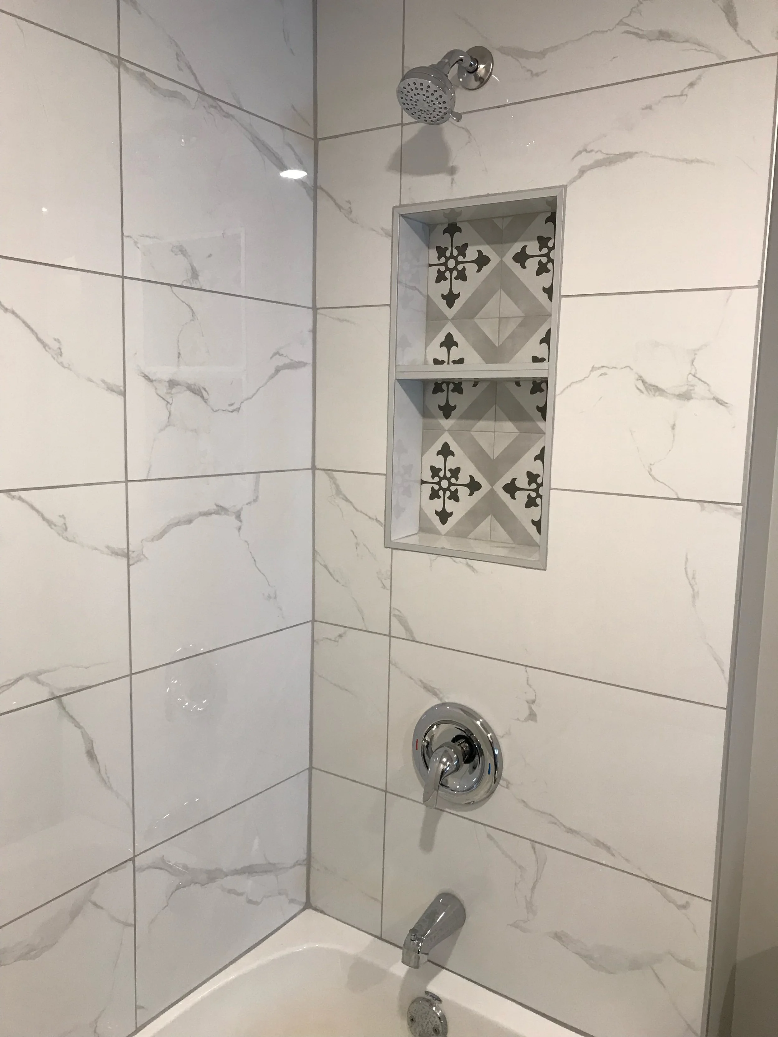 Shower with marble-patterned tiles, a showerhead, a recessed shelf with decorative tile backing, and a faucet.