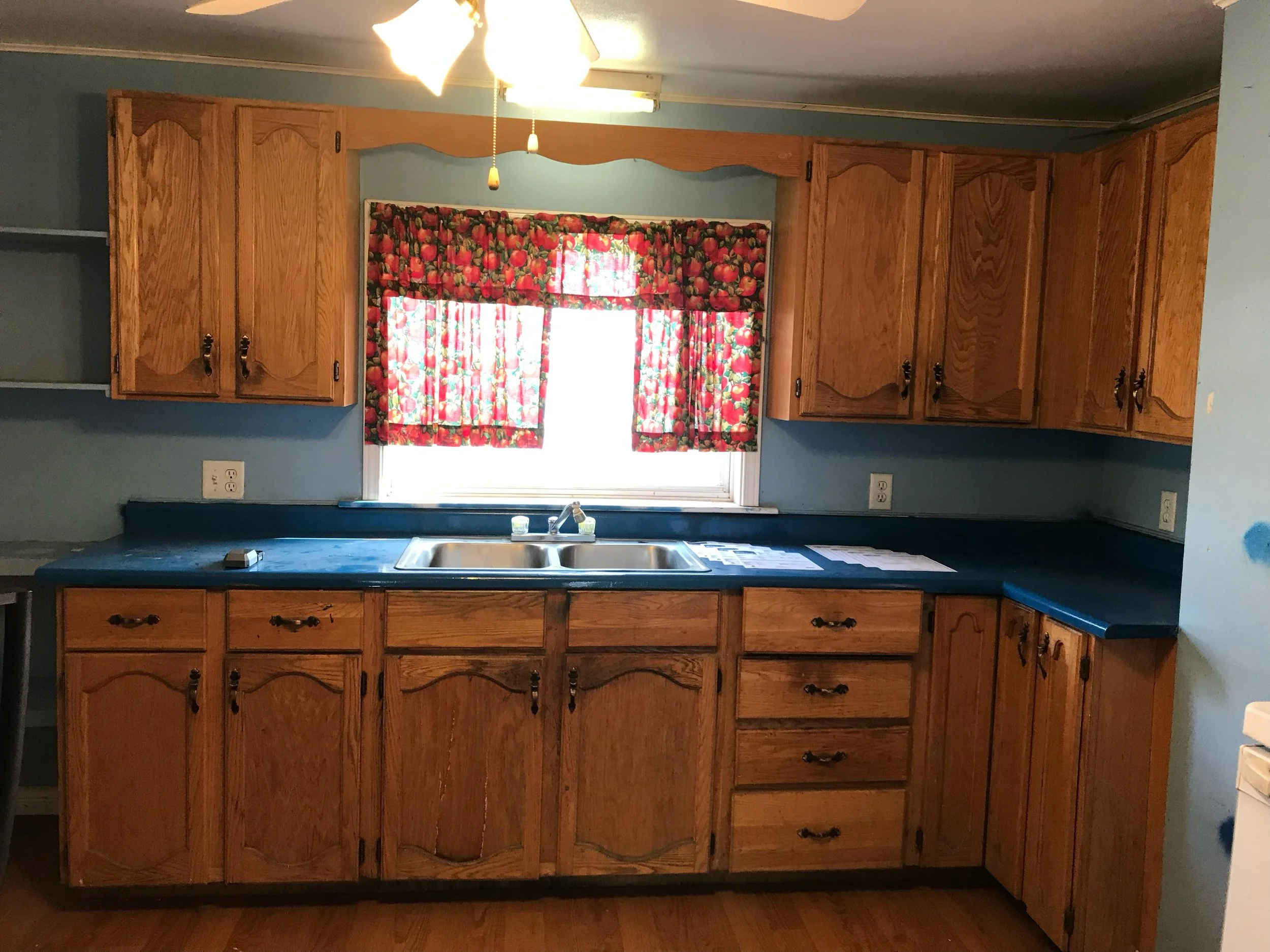 Wooden kitchen cabinets above and below a blue countertop with a double sink. A window with colorful strawberry-patterned curtains is above the sink, and there are some papers on the countertop.