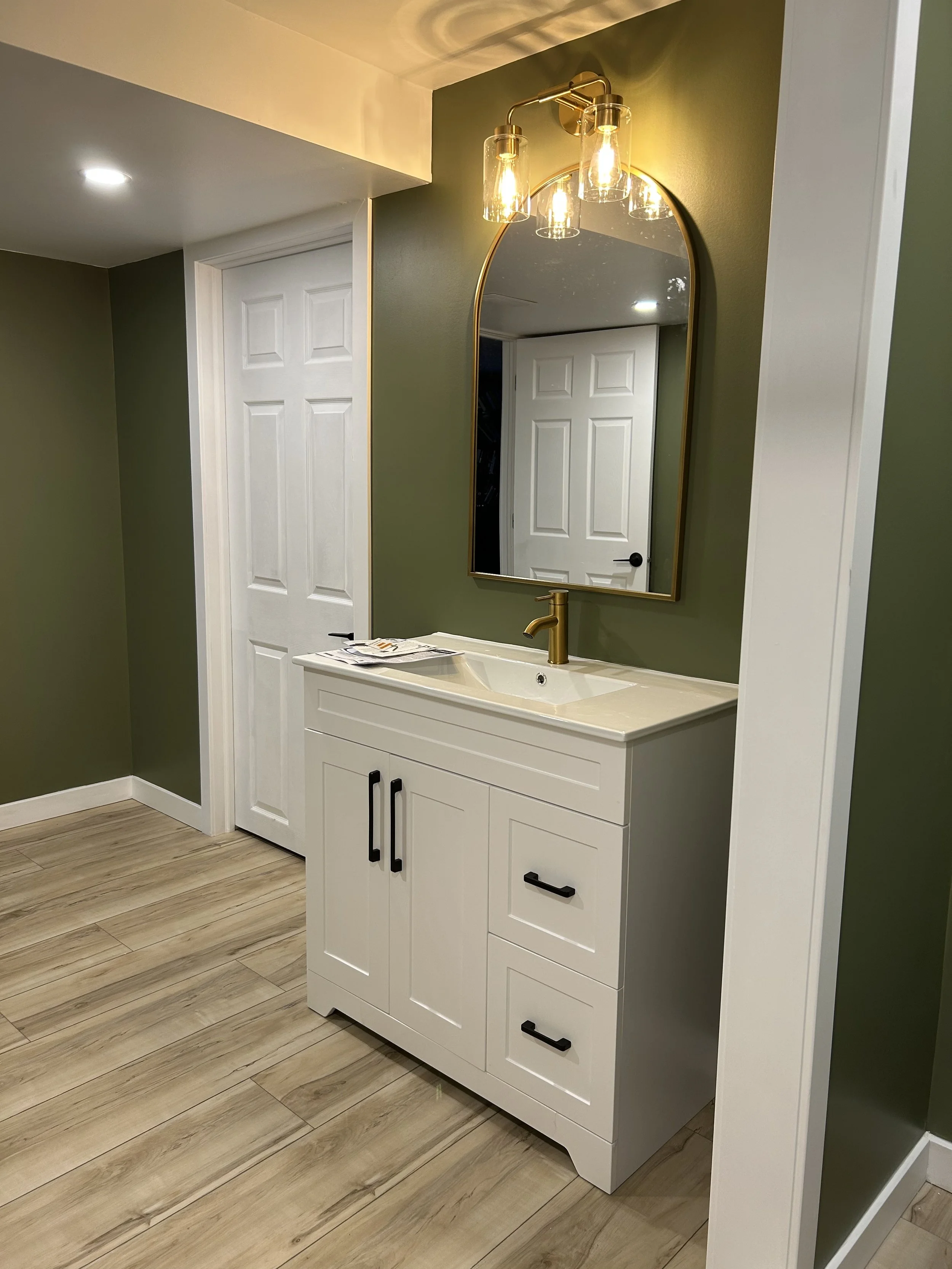 A modern bathroom vanity with a white cabinet, black handles, a beige countertop, a gold faucet, and an oval mirror with a gold frame. A three-light fixture with glass shades is mounted above the mirror. The bathroom has green and white walls, wooden floor, and a door in the background.