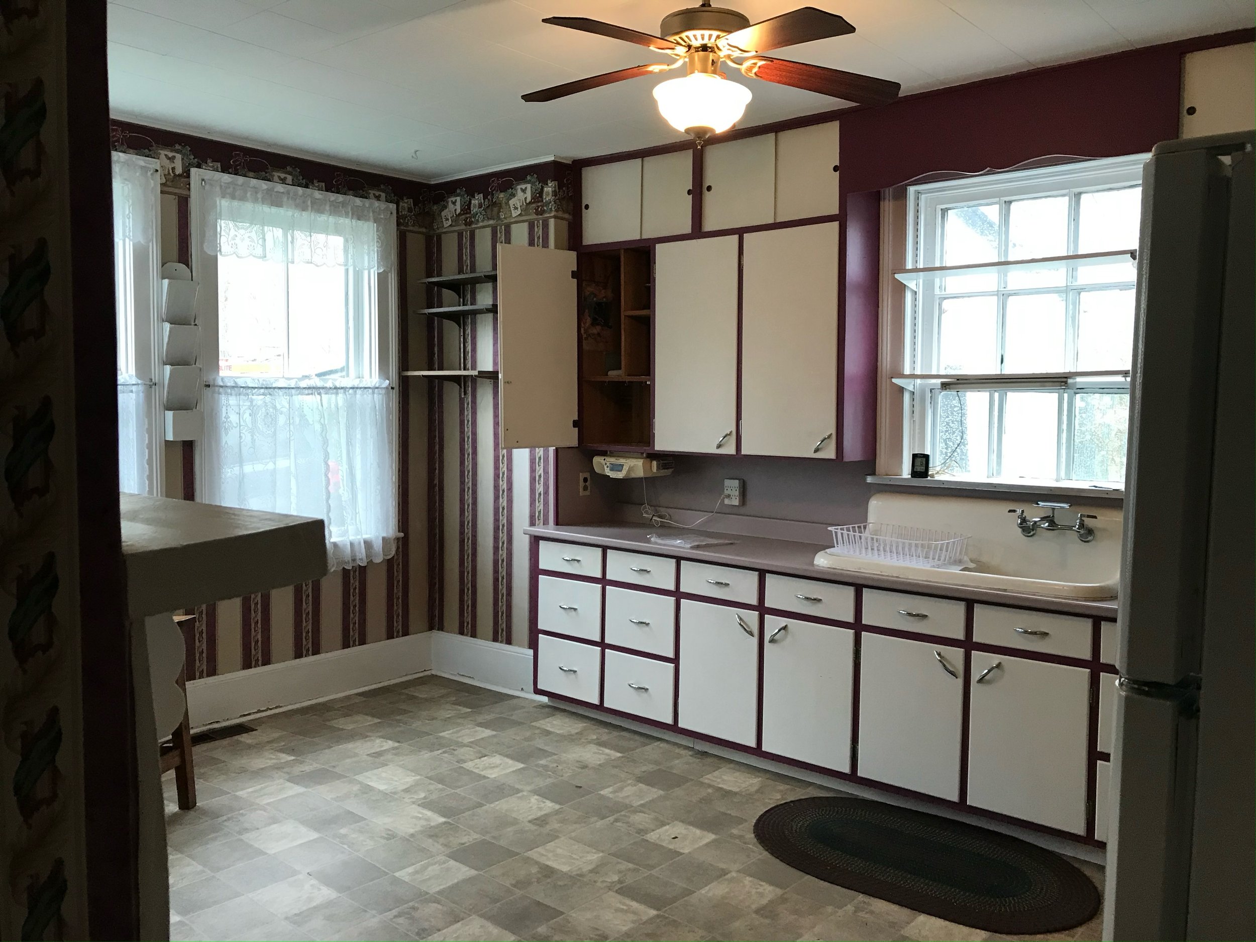 Kitchen with vintage decor, white cabinets with maroon trim, a double window above the sink, open cupboard, ceiling fan, and patterned wallpaper.
