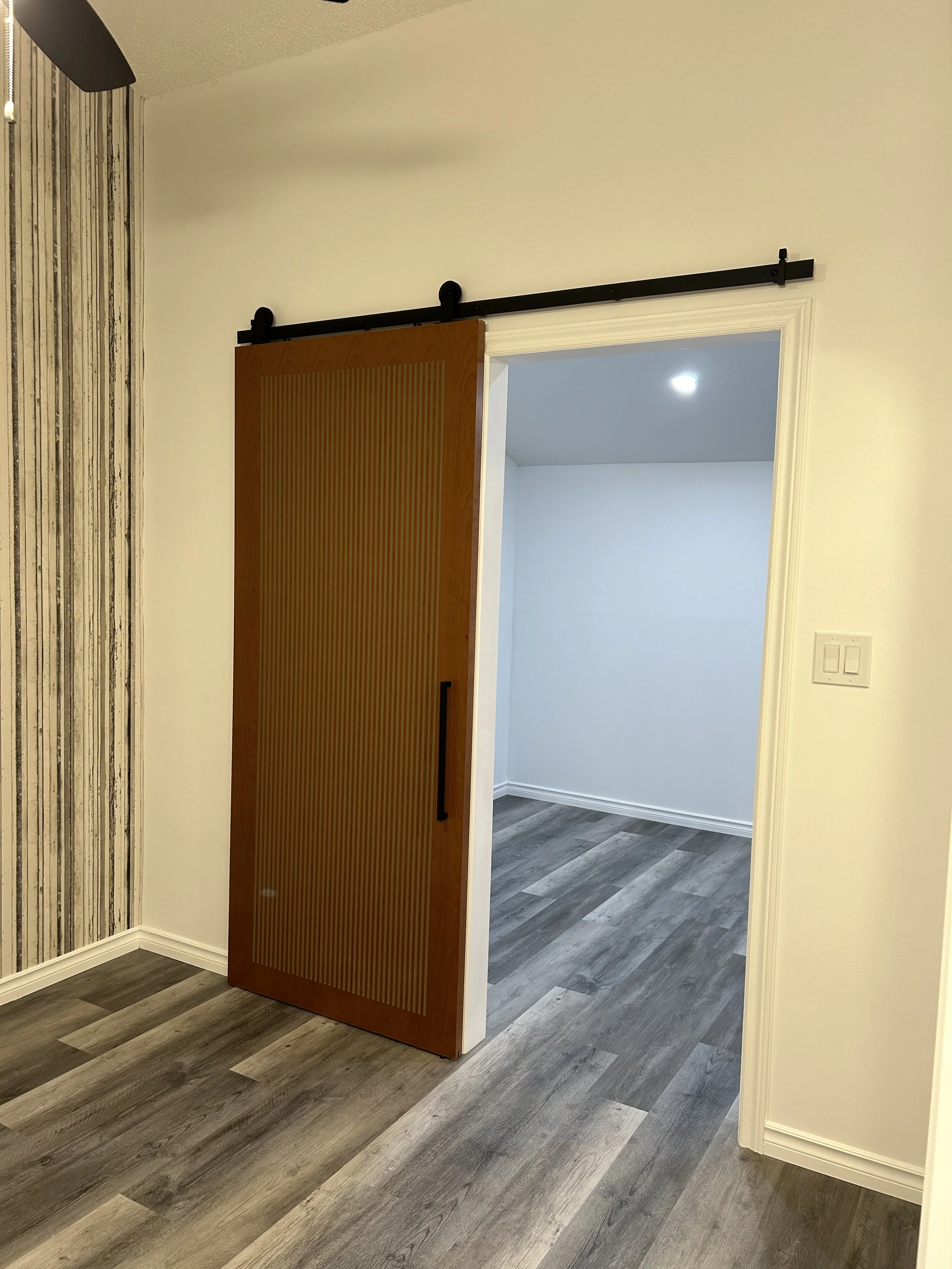 Interior view of a room with a barn door style sliding door, partially open, revealing an empty room with wood-look flooring and white walls.