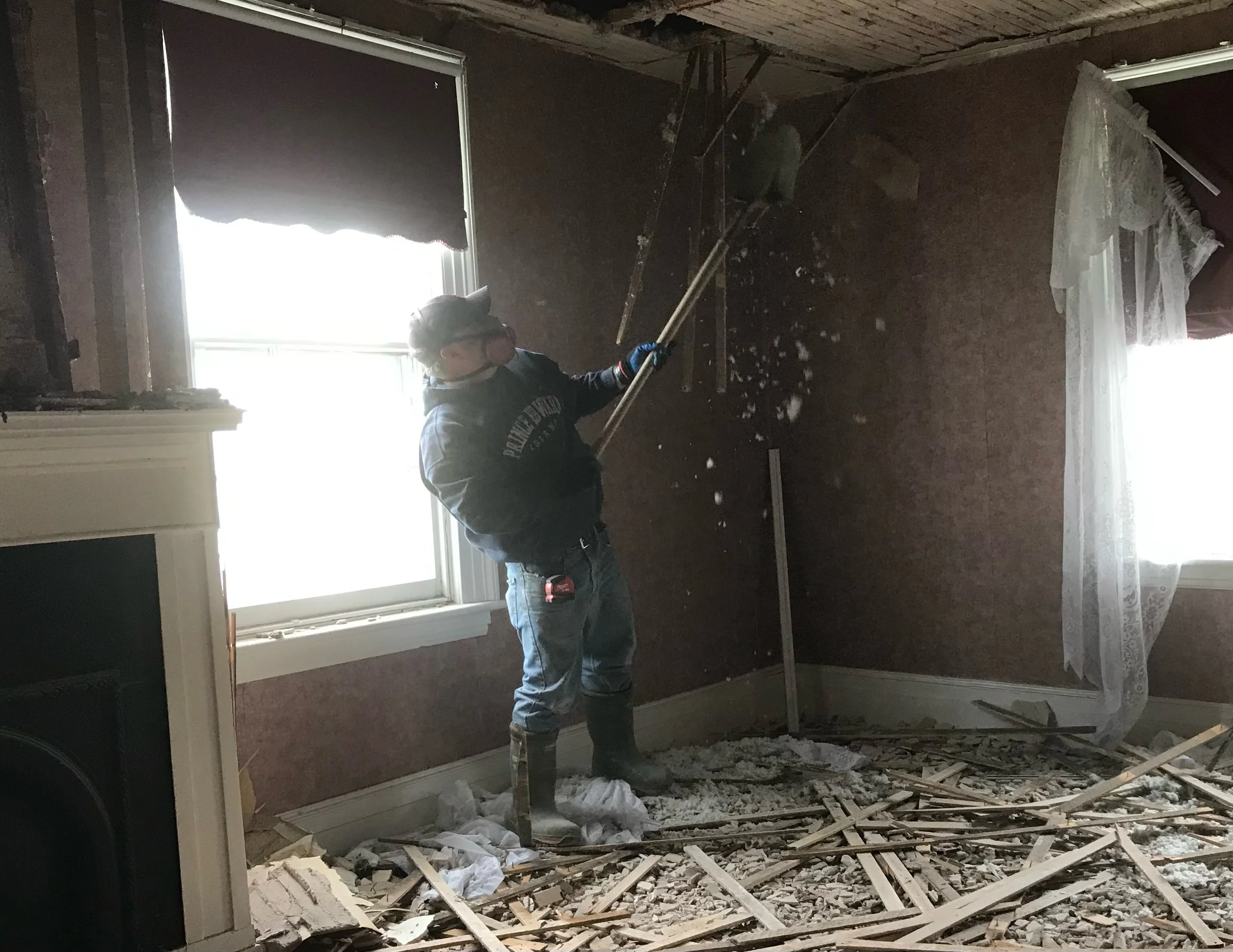 Person wearing safety glasses, gloves, and rubber boots removing debris from a room with a sledgehammer, exposed wooden floor framing, and curtains hanging by the window, indicating demolition activity.