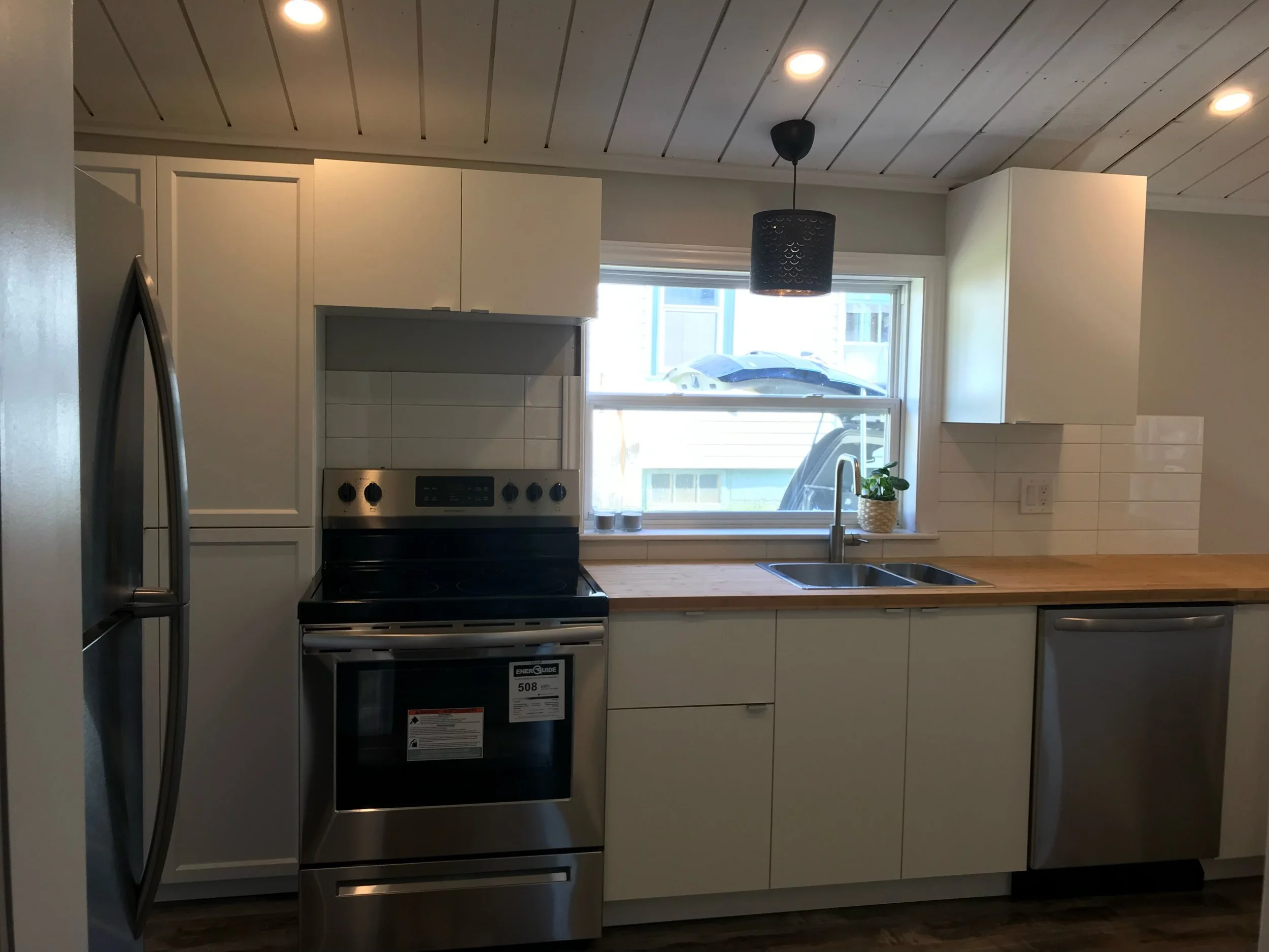 Kitchen with white cabinets, wooden countertop, stainless steel refrigerator, oven, and dishwasher, window with a mountain view, hanging light fixture, and a small plant on the windowsill.
