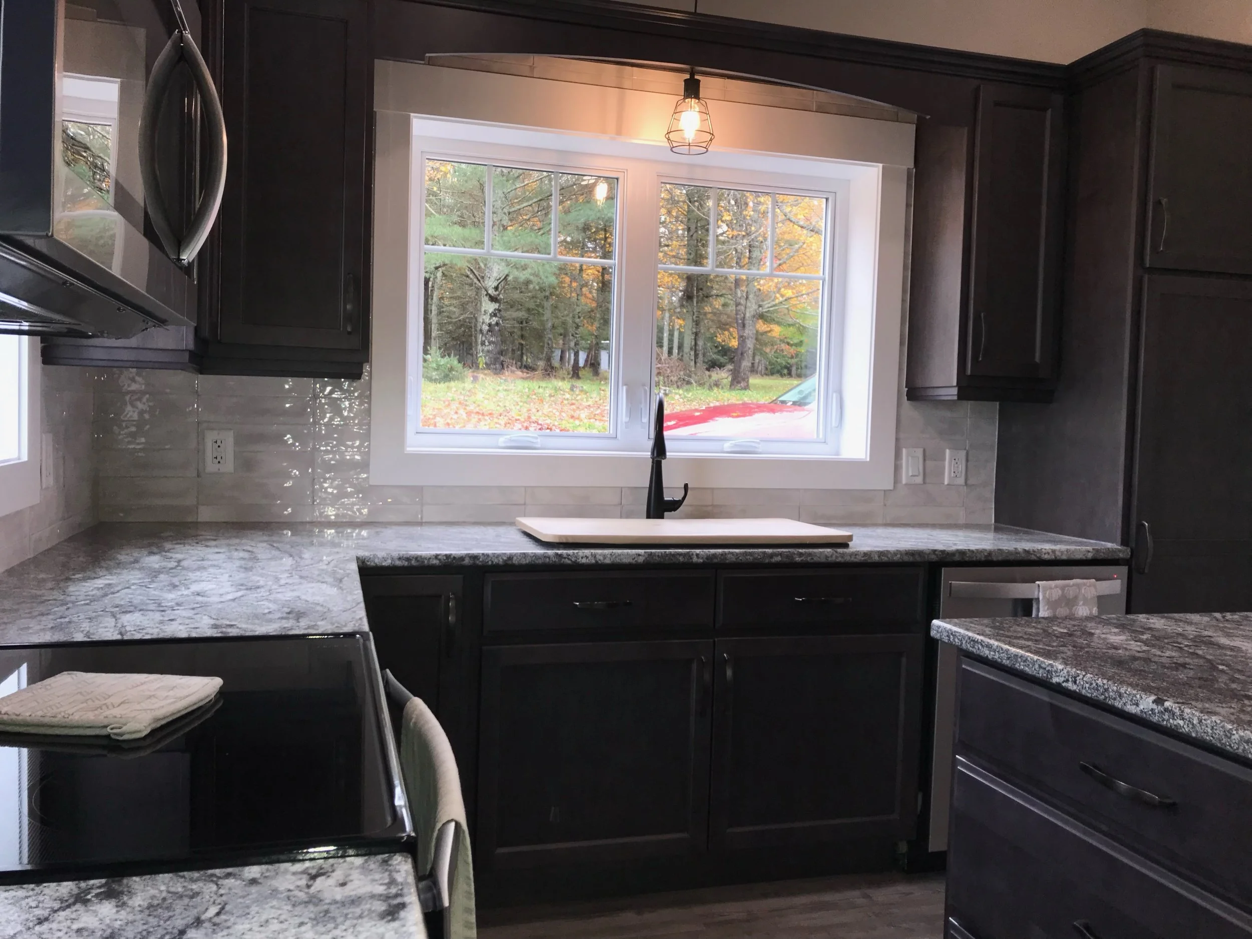 A modern kitchen with black cabinetry, granite countertops, a white sink under a large window, and a view of trees outside. There is a black oven with a towel hanging on its handle and a hanging light fixture above the sink.