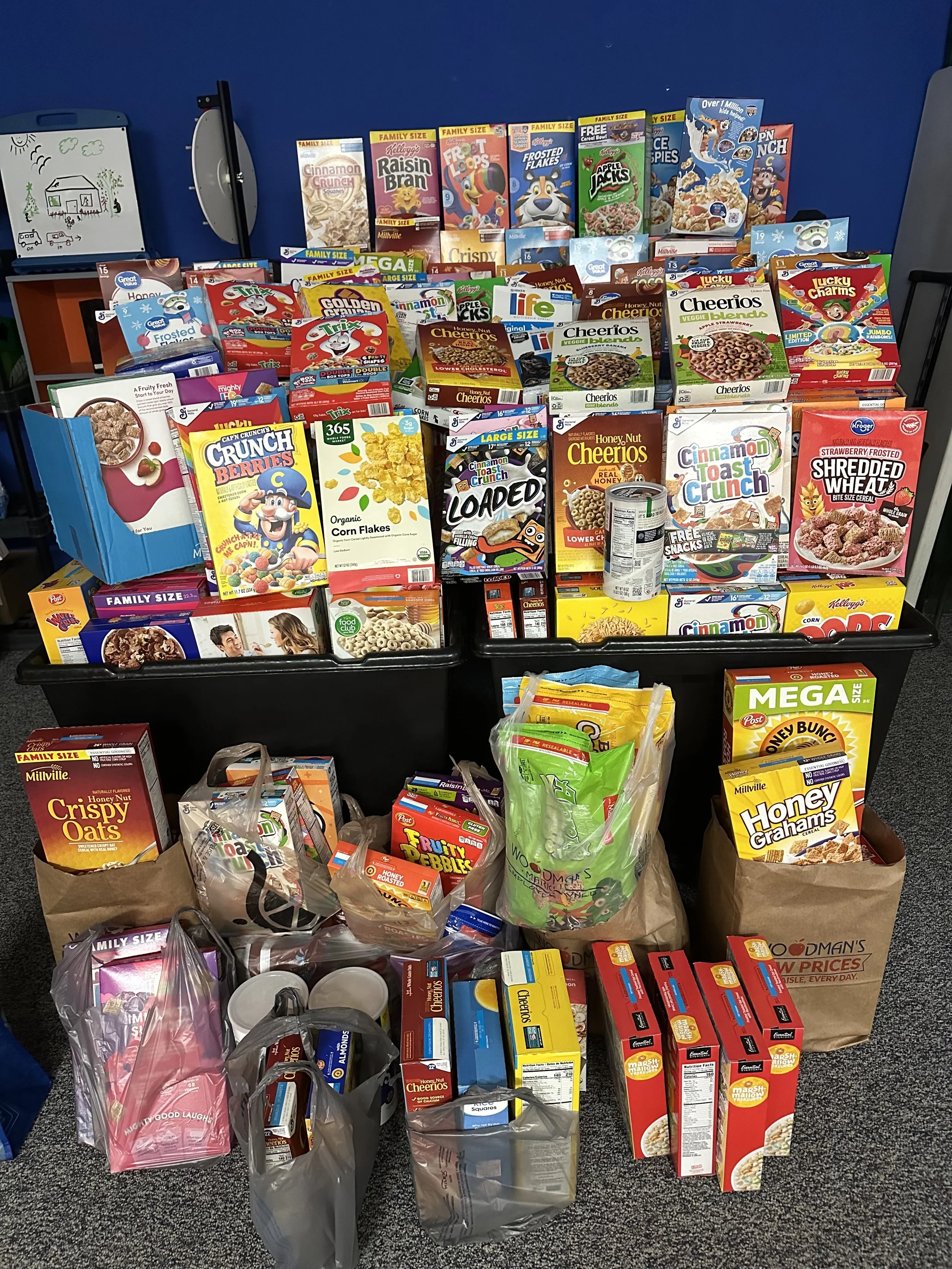 A large collection of cereal boxes and packaged breakfast foods on display, including Cheerios, Cinnamon Toast Crunch, Lucky Charms, Frosted Cheerios, and more, with bags of additional grocery items and food packages in front.