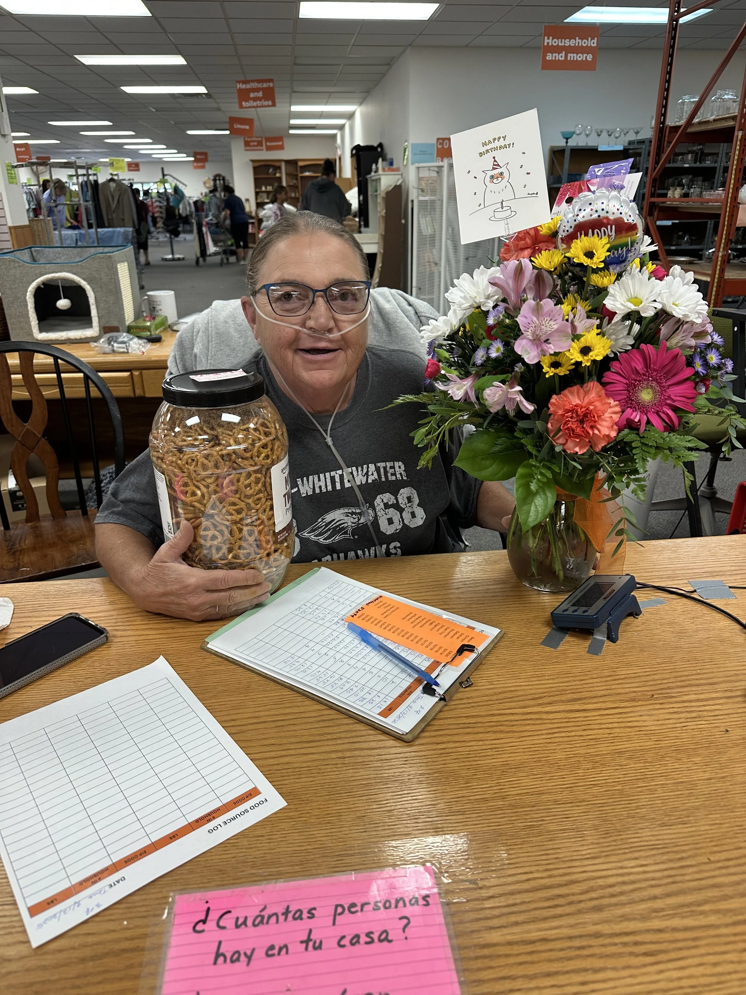 A woman with a nasal cannula holding a large jar of pretzels is sitting at a table with a colorful bouquet of flowers and a birthday card that says 'Happy Birthday!' in a store or community center. The table has papers, a pen, and a small electronic device. There are other people browsing in the background.
