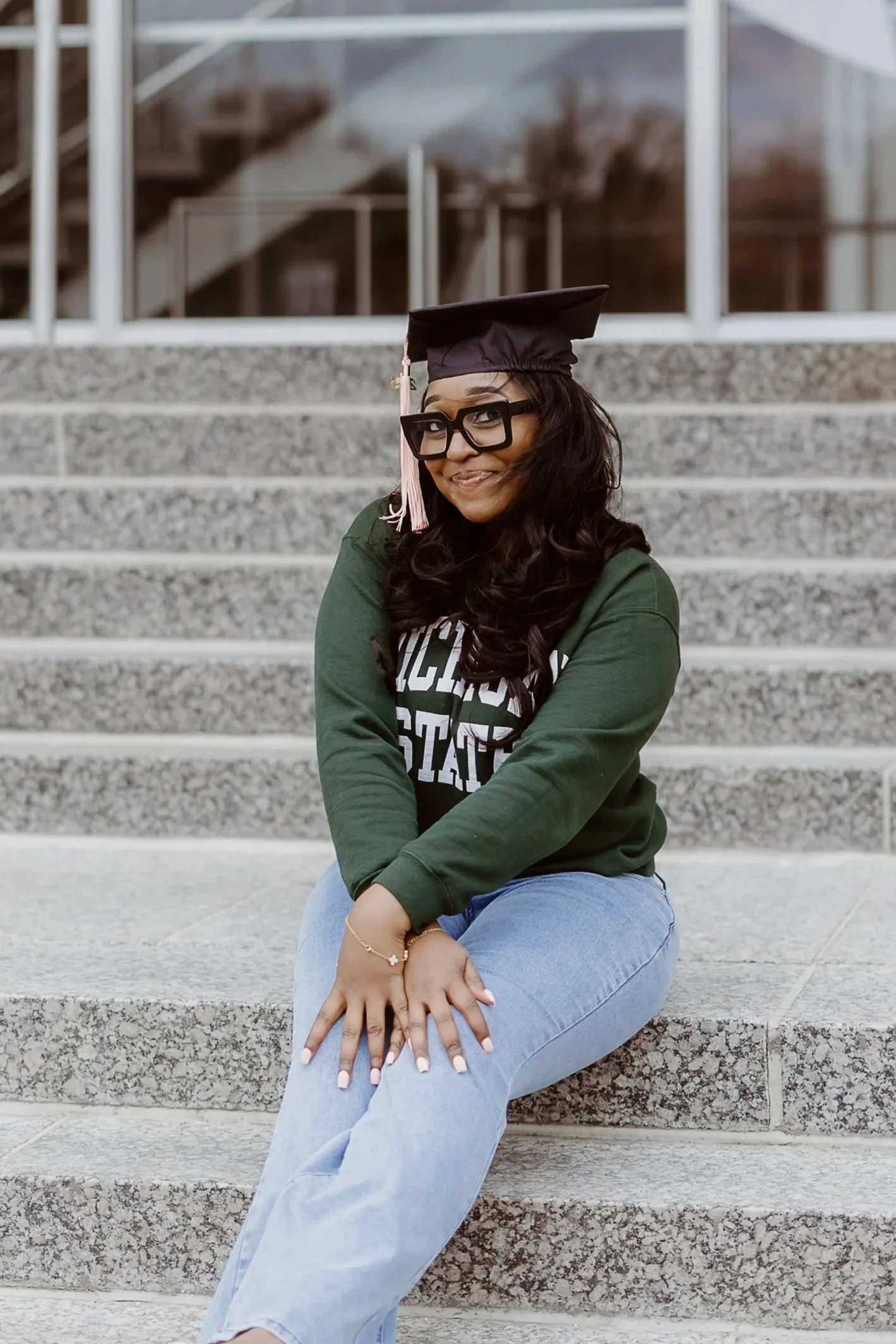 Woman in graduation cap and gown sitting on outdoor stairs, smiling, with glasses and dark wavy hair.