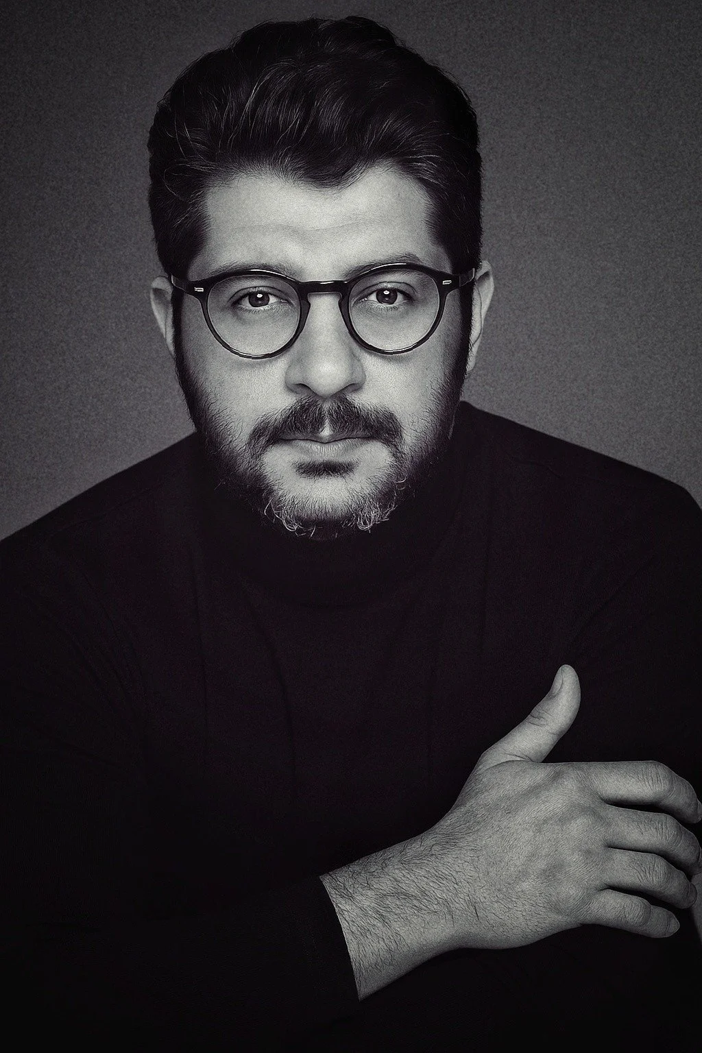 A black and white portrait of a man with glasses and a beard, wearing a black turtleneck, looking directly at the camera with a serious expression.