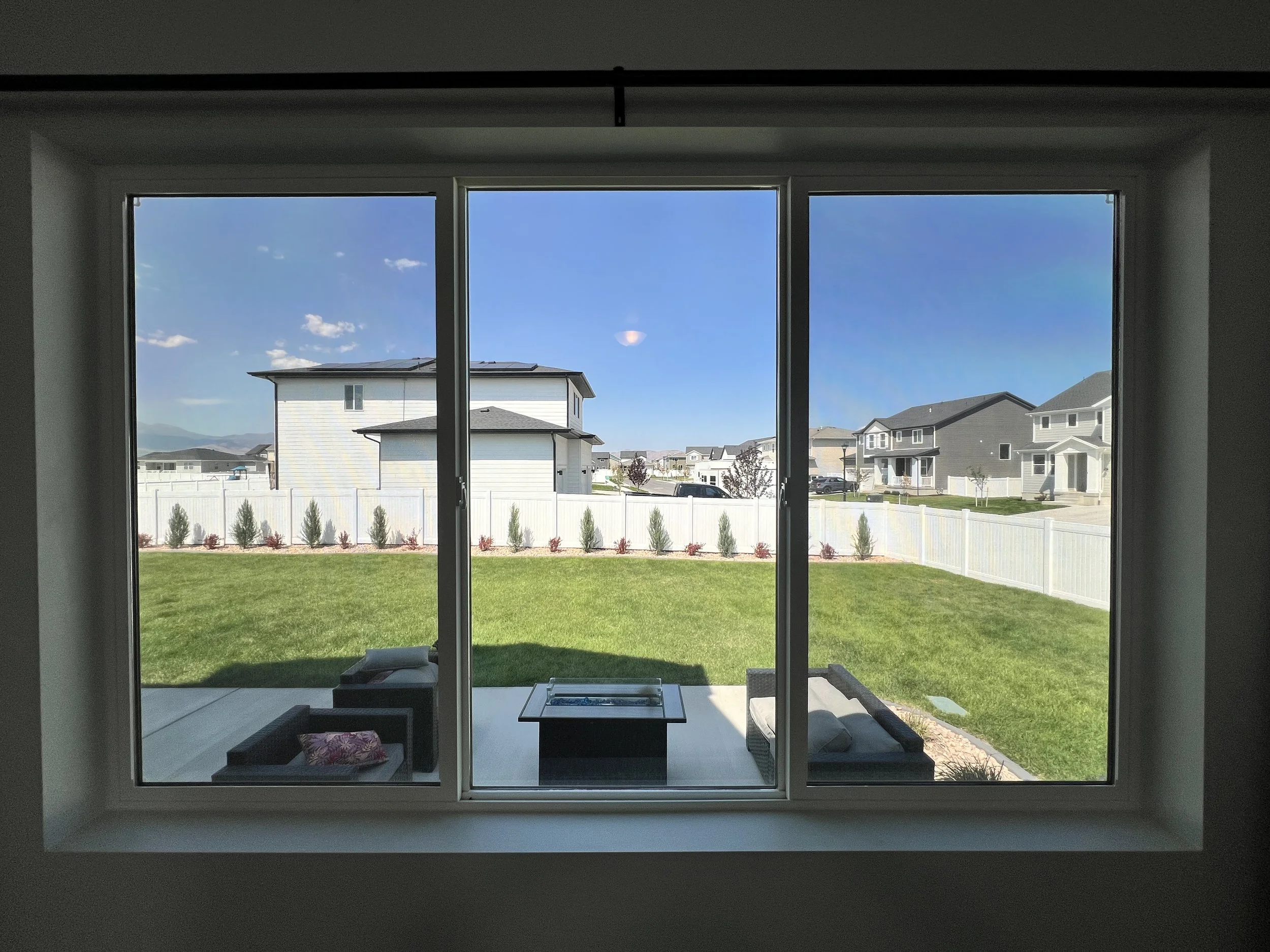 View through a large window showing a backyard with green grass, outdoor furniture, a white fence, and neighboring houses under a blue sky.
