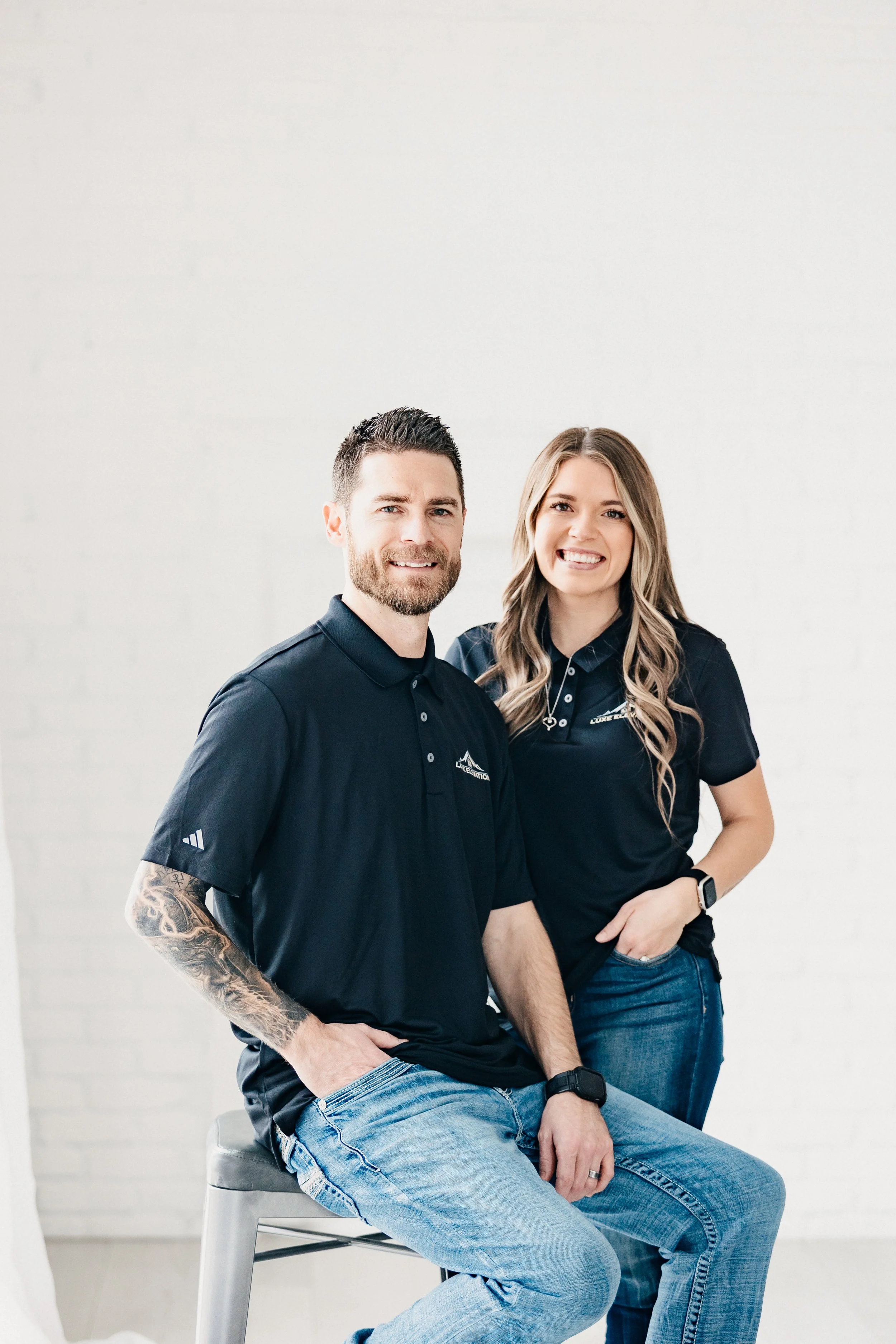 A man and woman smiling and posing together against a white background, wearing matching dark blue polo shirts with logos, with the man sitting on a stool and the woman standing.