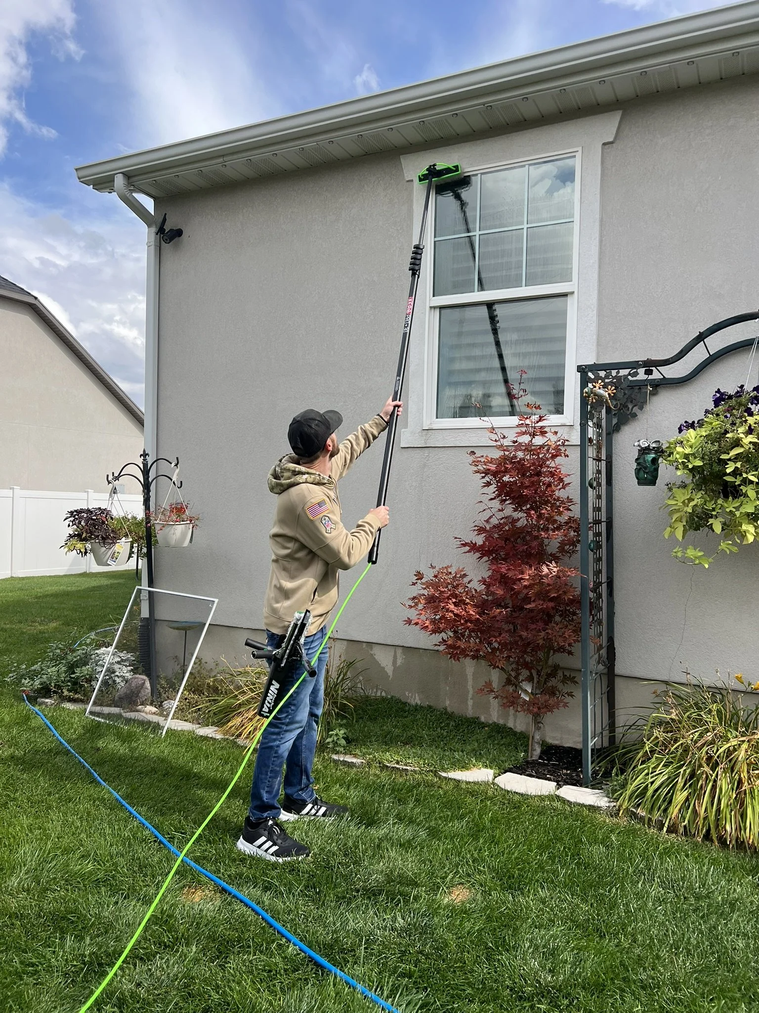 A person using a long-handled cleaning tool to clean a window on the second floor of a house. The person is standing on a manicured lawn, and there are plants and a decorative trellis nearby. The sky is partly cloudy.