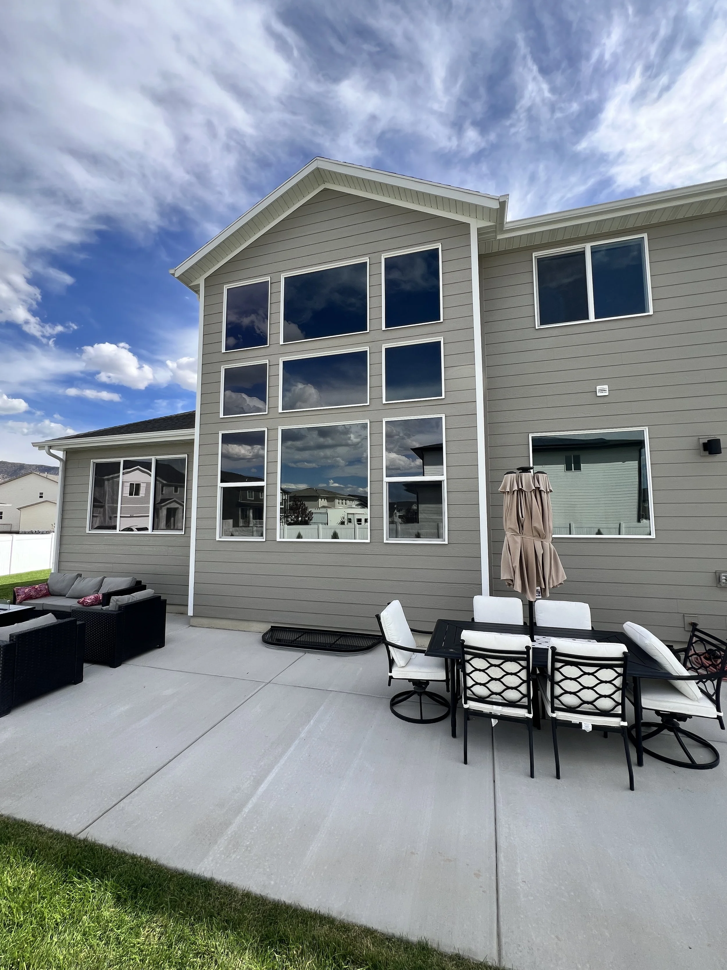 Backyard patio with outdoor furniture including a black wicker sofa and a black dining table with white chairs, a closed umbrella, and a modern house with large windows reflecting the blue sky and clouds.