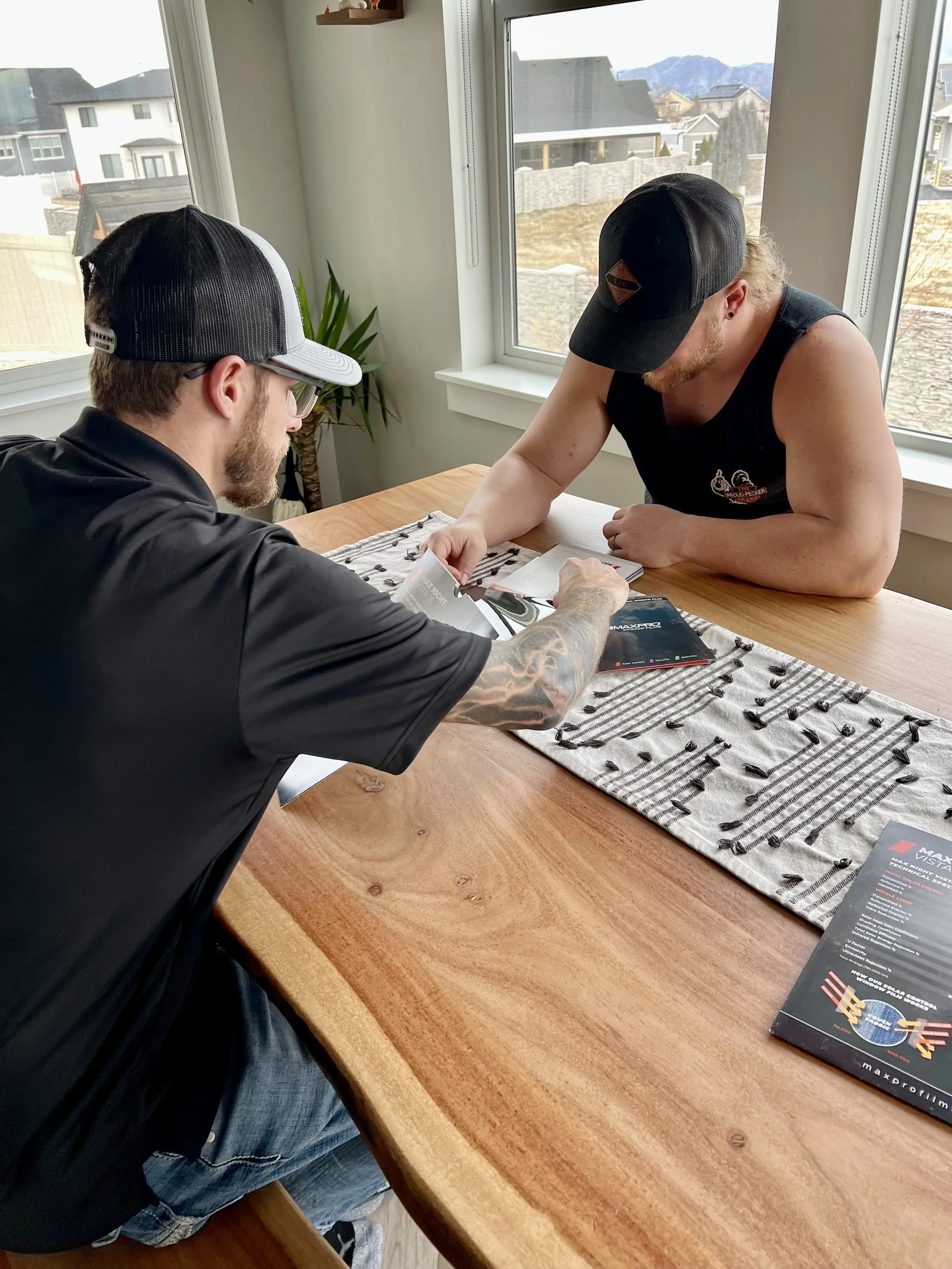 Two men sitting at a wooden table inside a house, looking at brochures or magazines, with large windows showing a neighborhood and mountains outside.