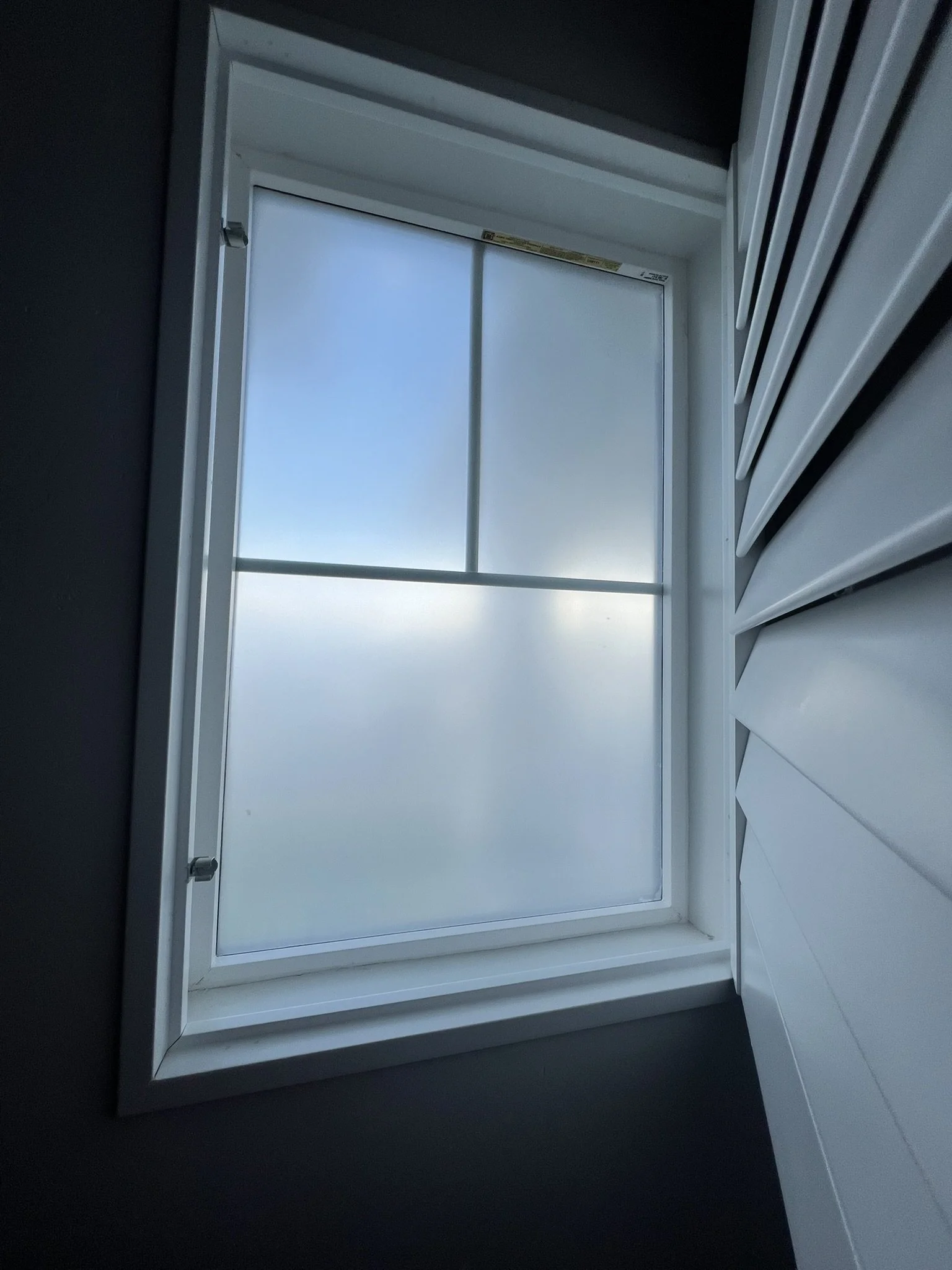 Close-up view of a white-framed window with frosted glass, set into a dark wall, with part of a white siding or paneling on the right side.