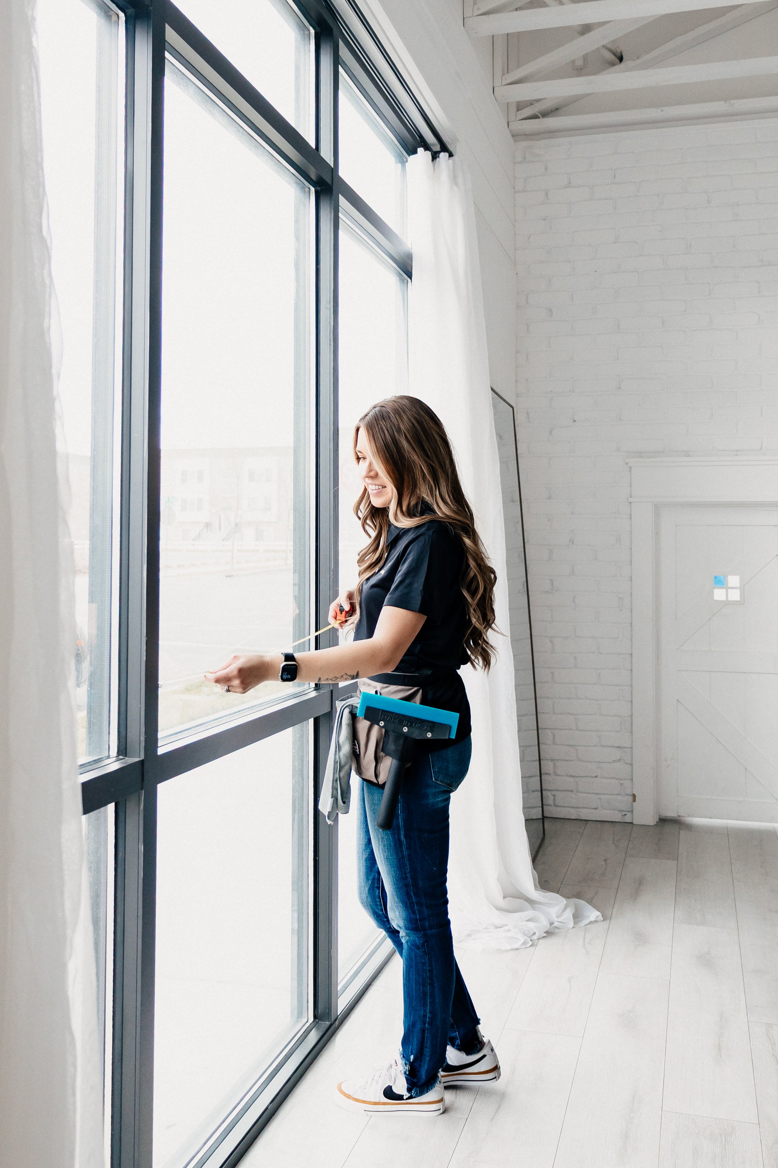 A smiling woman cleaning a large window inside a modern room with white brick walls, white curtains, and light wood flooring.