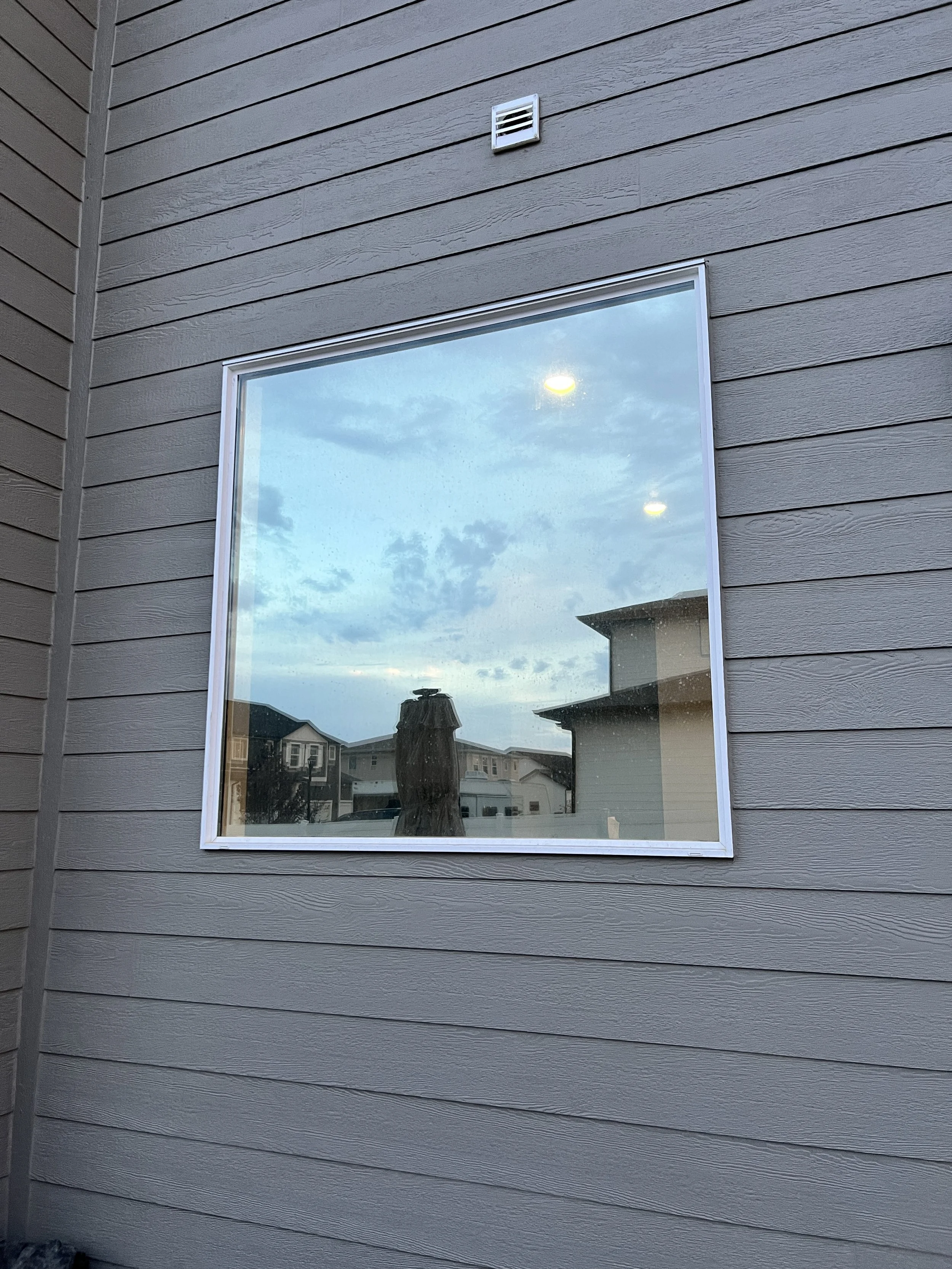 A residential house with gray wooden siding and a large window reflecting a blue sky with clouds and neighboring houses.