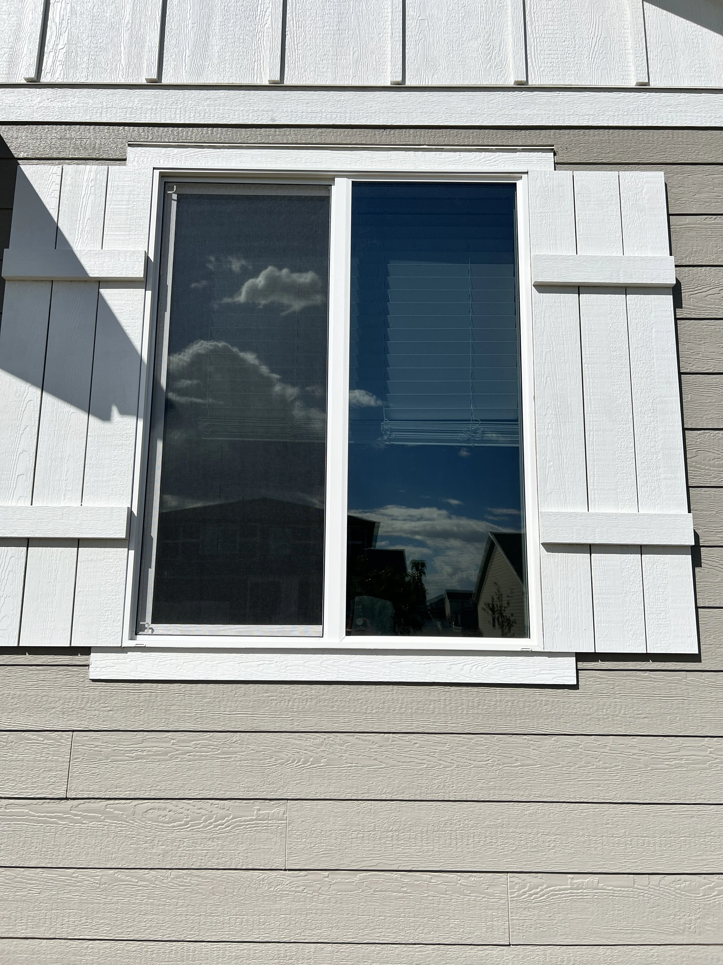 A house window reflecting a cloudy sky and neighboring houses, framed by white siding with decorative shutters.
