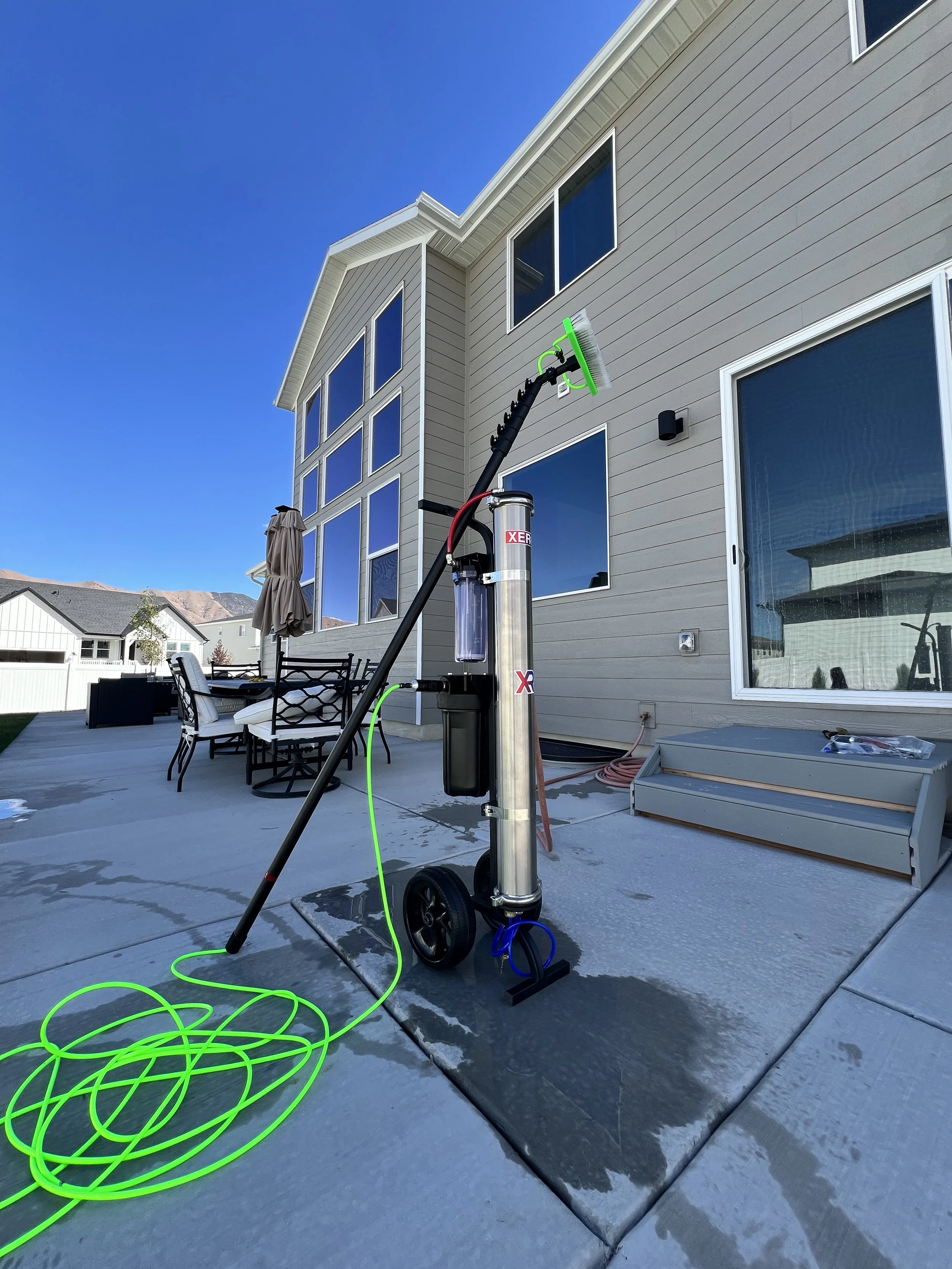 A house exterior with a pressure washer on a patio, connected to a bright green hose, and outdoor furniture including a table and chairs under an umbrella.