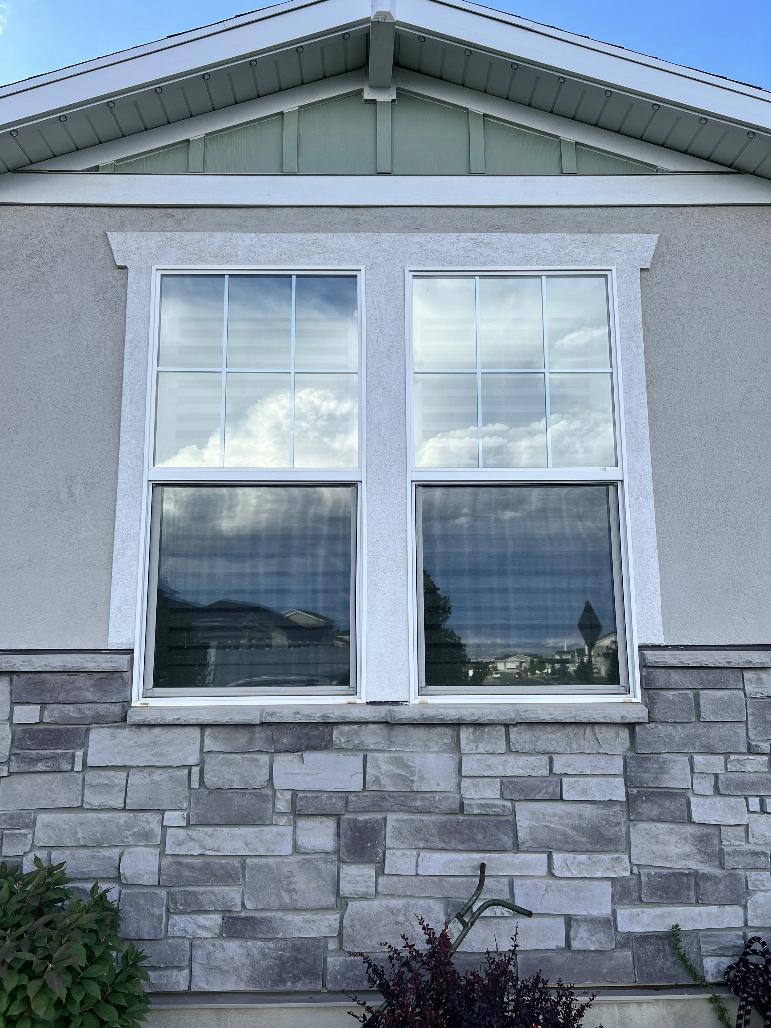 The front of a house with a stone foundation, two large windows reflecting a cloudy sky, and gray siding with a gable roof.
