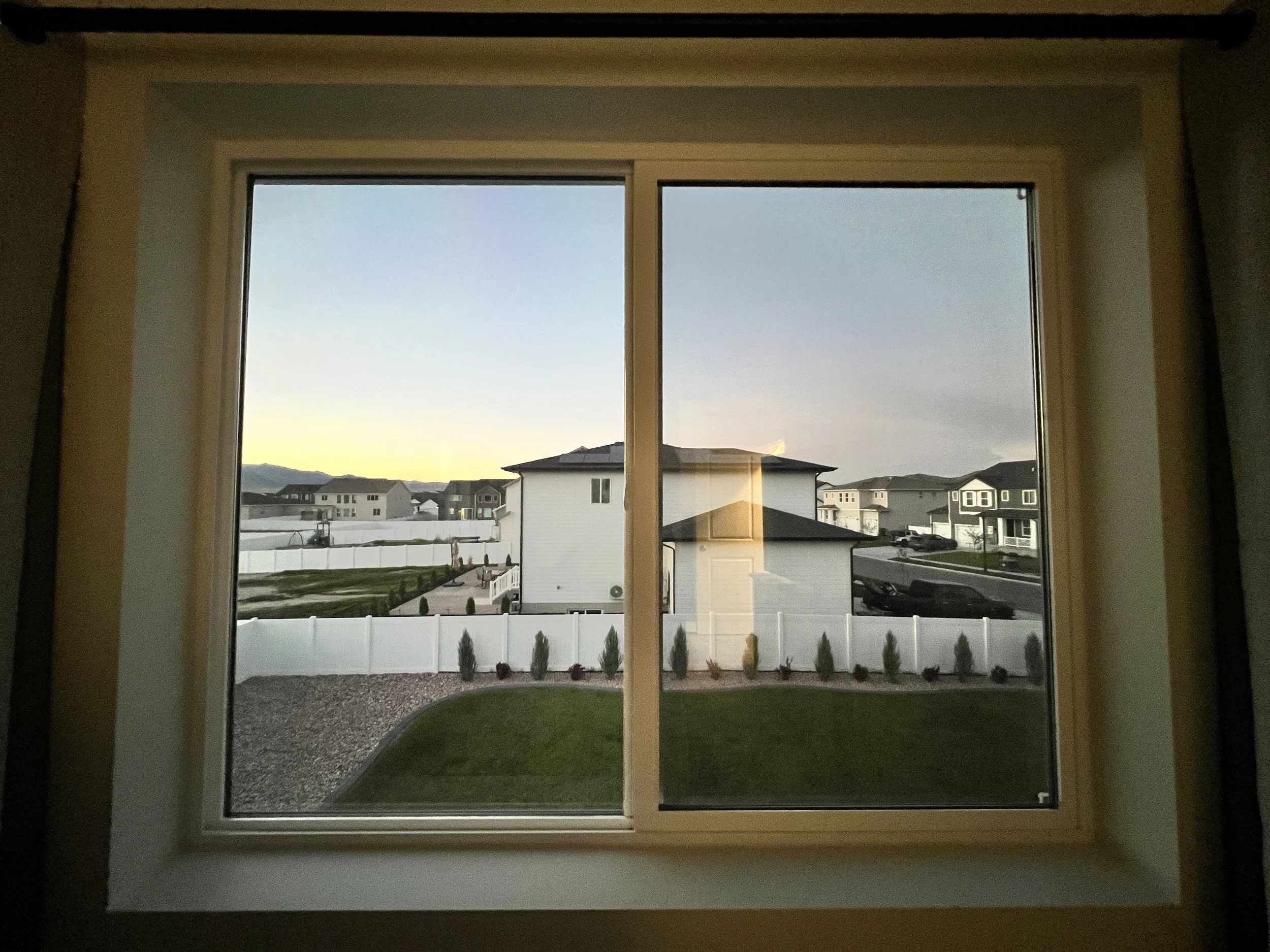 View of a suburban neighborhood through a large double-pane window at sunset, with white houses, a white picket fence, and a green lawn.