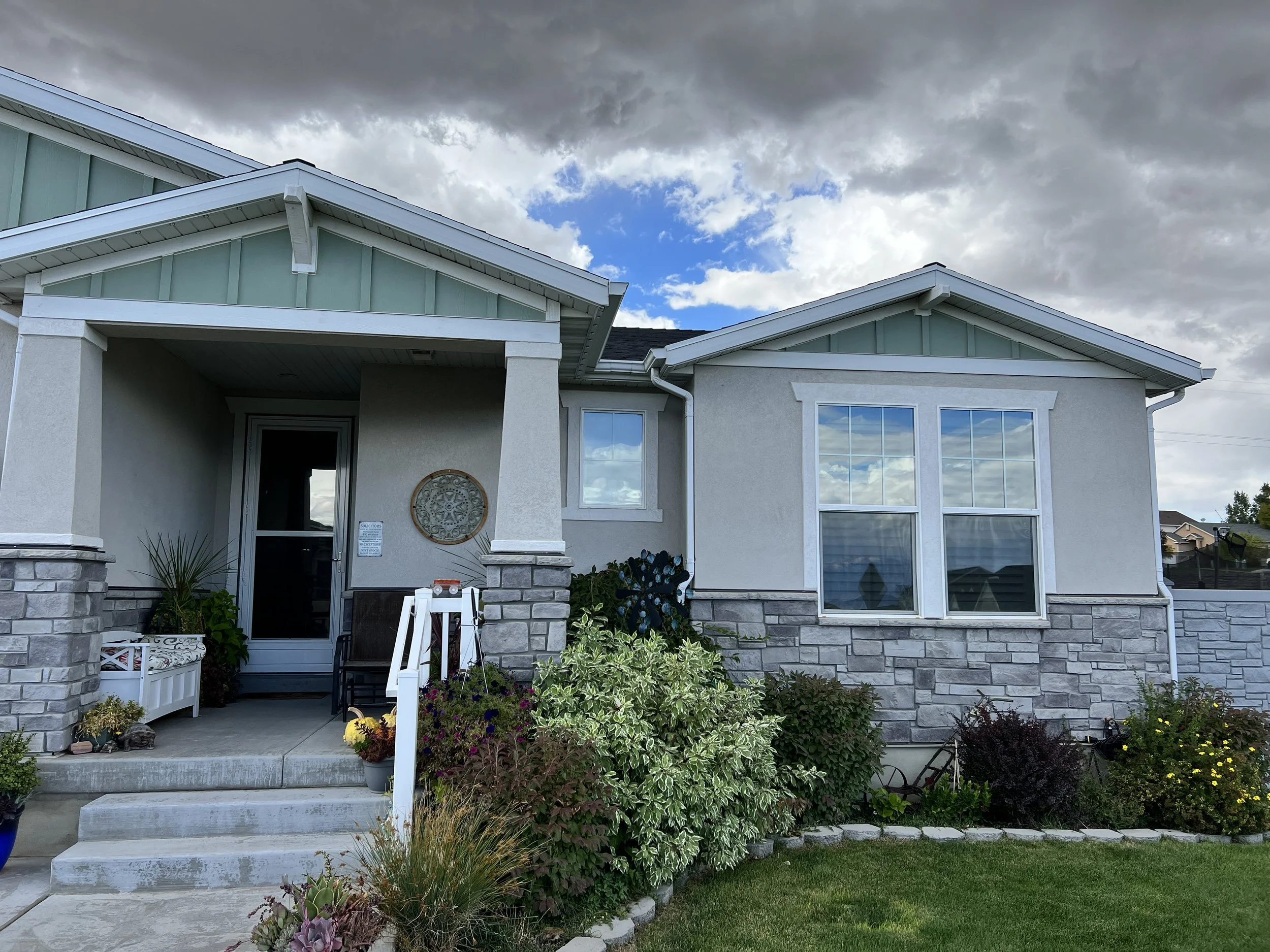 Front view of a modern house with stone and stucco exterior, large double-pane windows, and a small porch with plants and steps leading up.