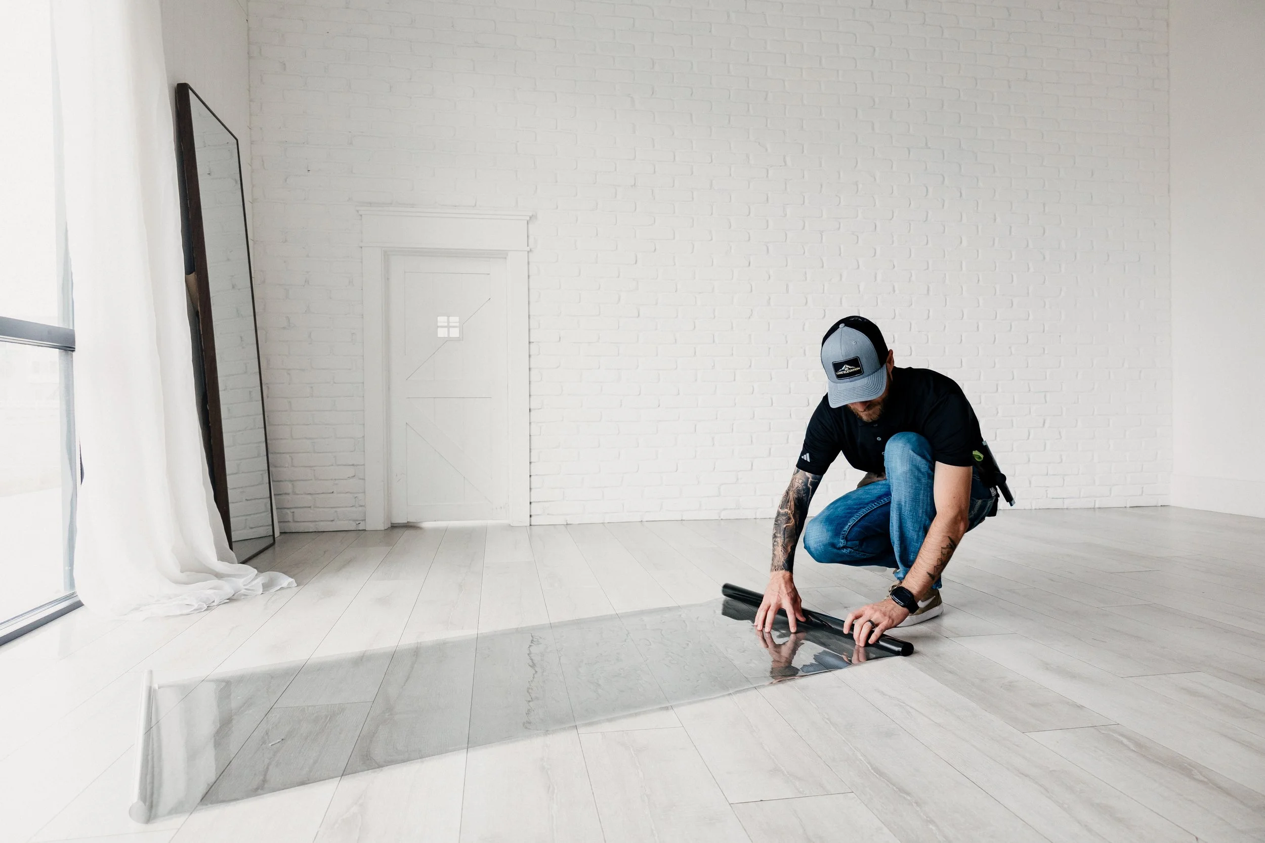 A man wearing a cap and black shirt is kneeling on a light-colored wooden floor, unrolling a roll of clear adhesive film or vinyl near a white brick wall, with a mirror and window with white curtains to the side.